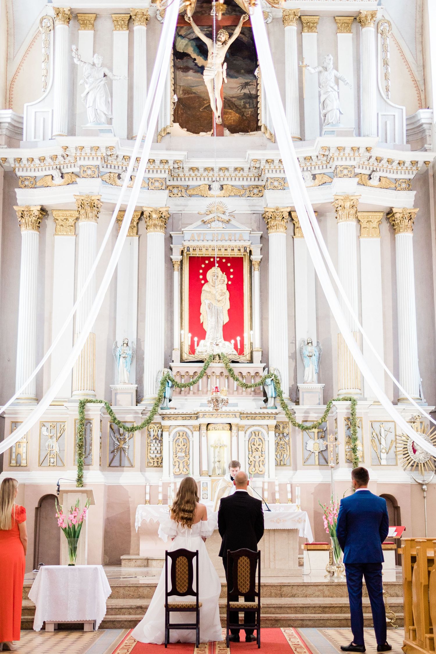 Interior view of an ornate white church with gold accents and chandeliers showing people seated in wooden pews.