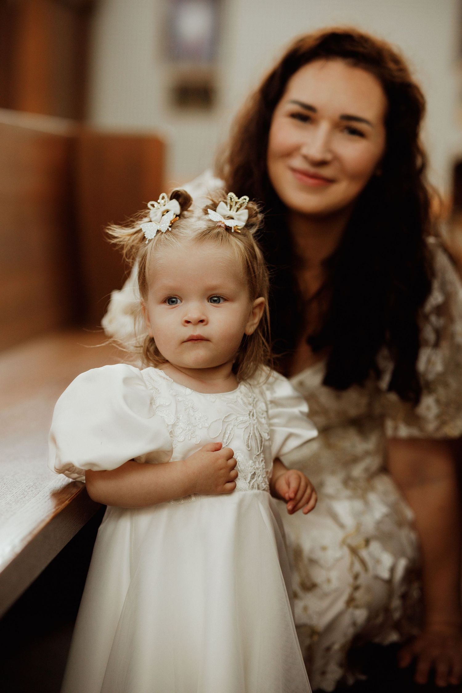 A toddler in a white lace dress with small flowers in her hair stands indoors during a formal occasion.