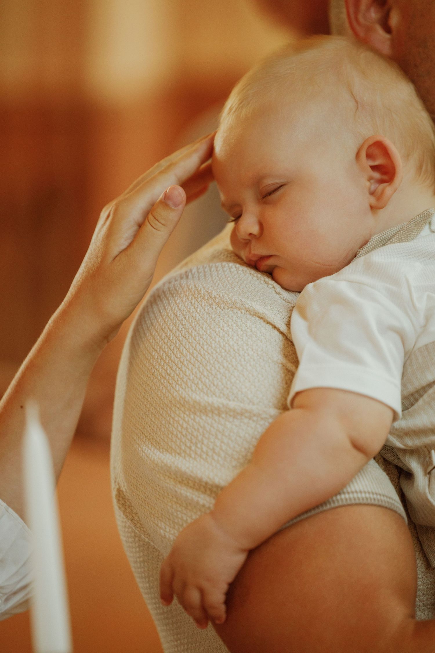 Sleeping baby rests peacefully against caregiver in warm indoor lighting.