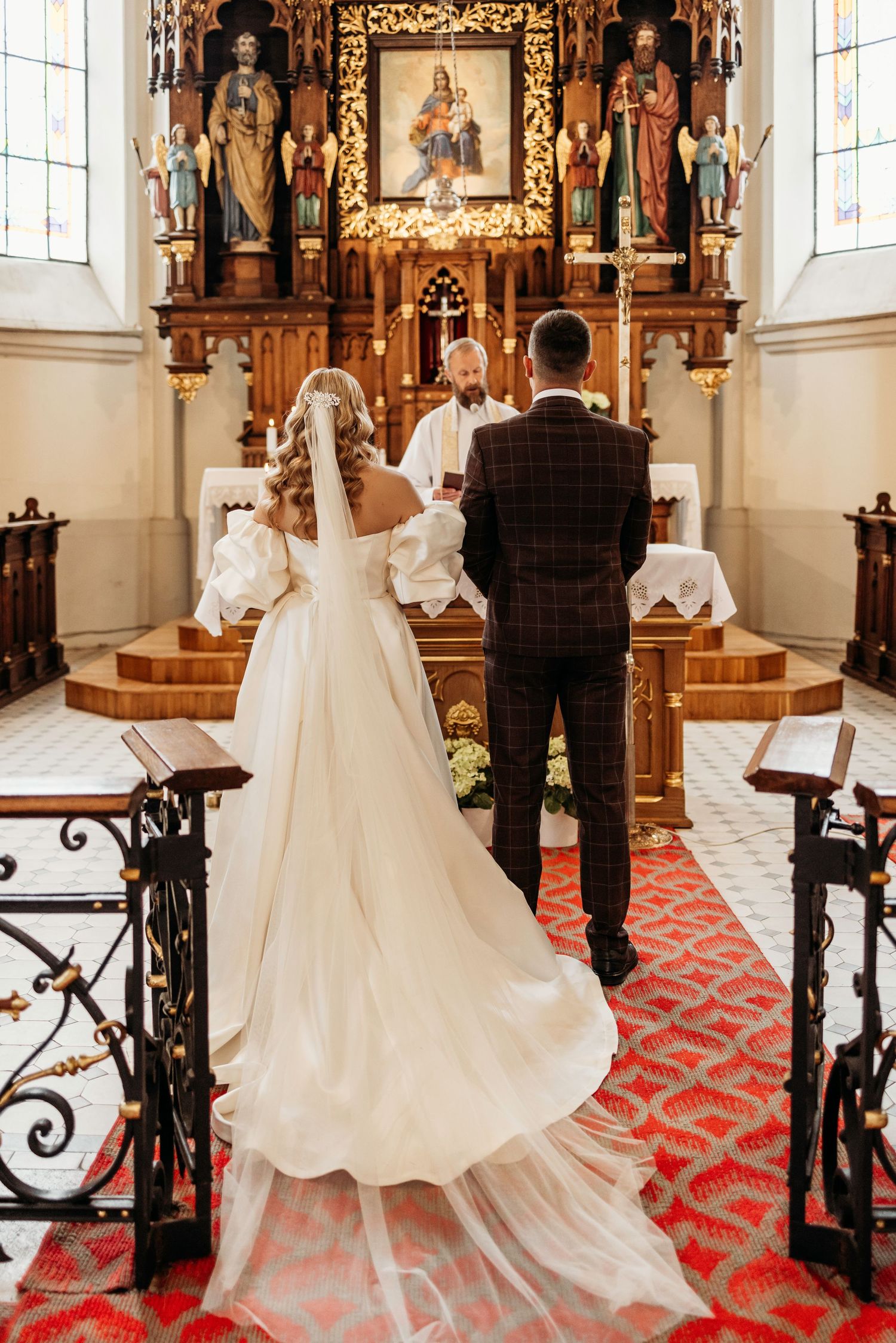 A wedding ceremony takes place inside an ornate church with gothic architecture and a red carpeted aisle.