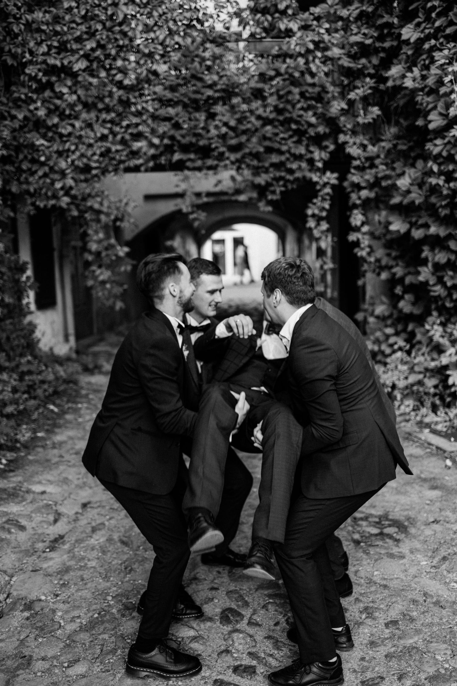 Three groomsmen in black suits playfully lift and support each other in a candid moment outdoors.