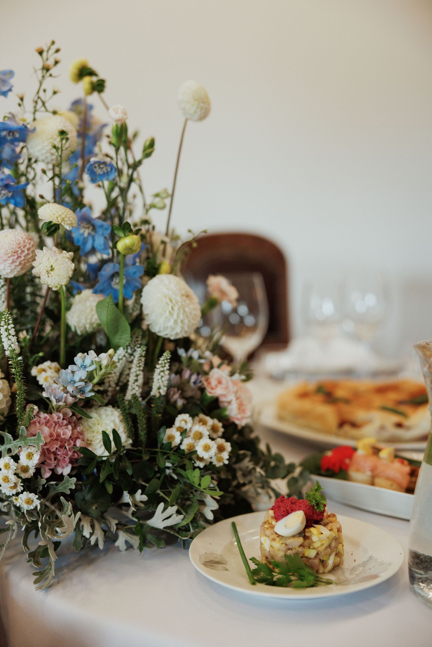 Garden-inspired centerpiece with blue delphiniums and white blooms beside plated appetizers.