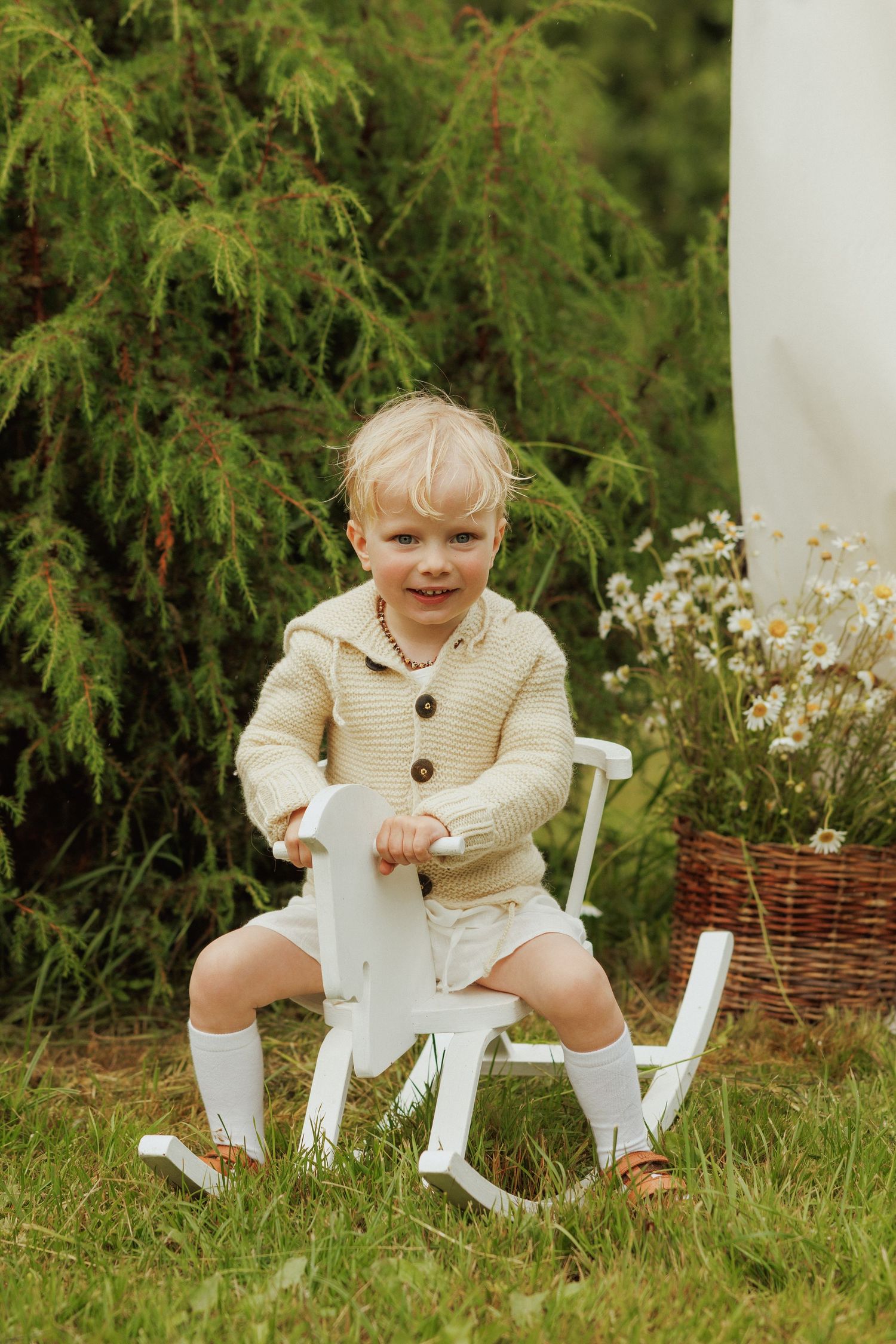 Smiling blonde toddler in beige knit sweater sitting on white rocking horse outdoors in garden.