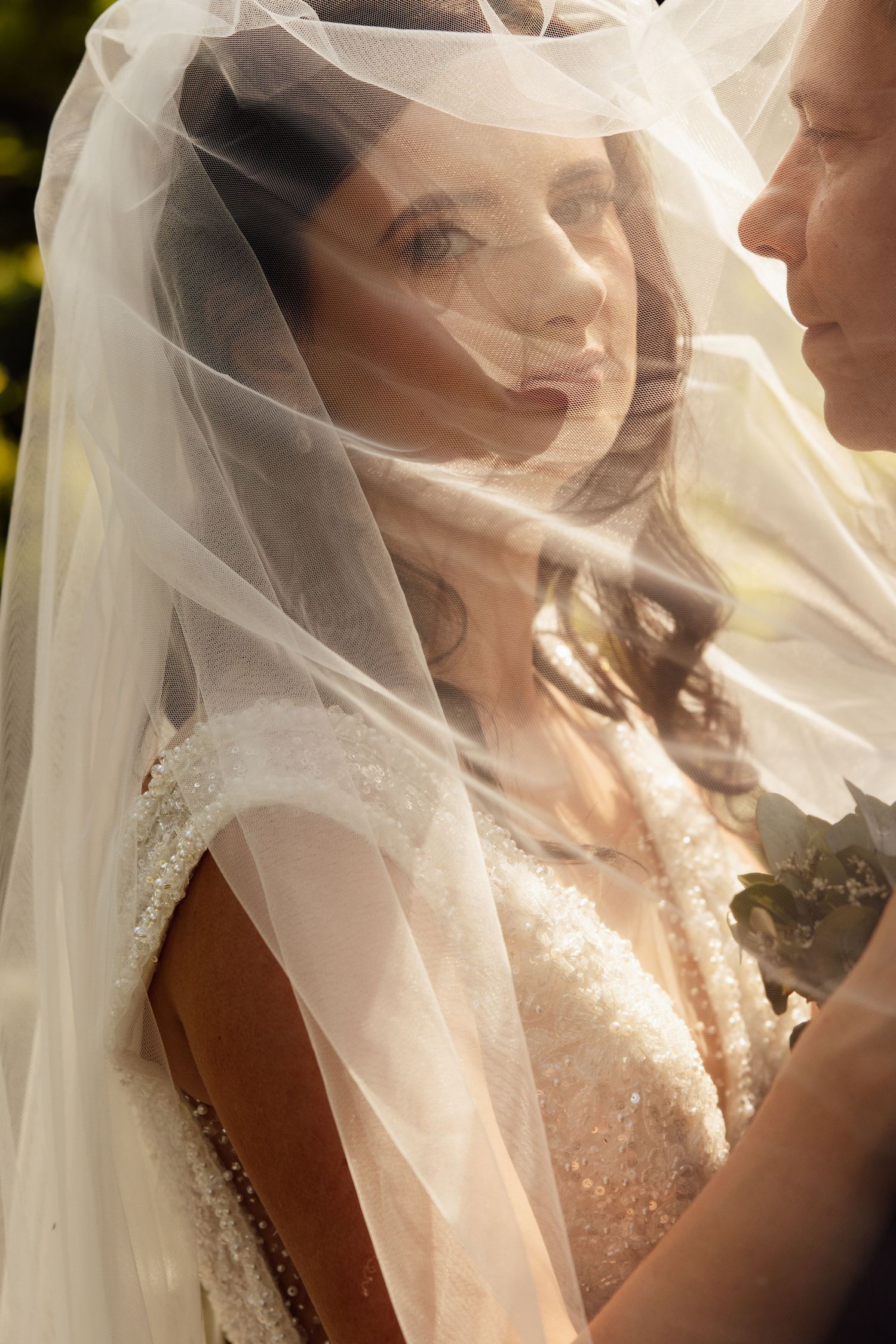 Bride in beaded gown and flowing veil gazes softly at groom during intimate wedding ceremony moment.