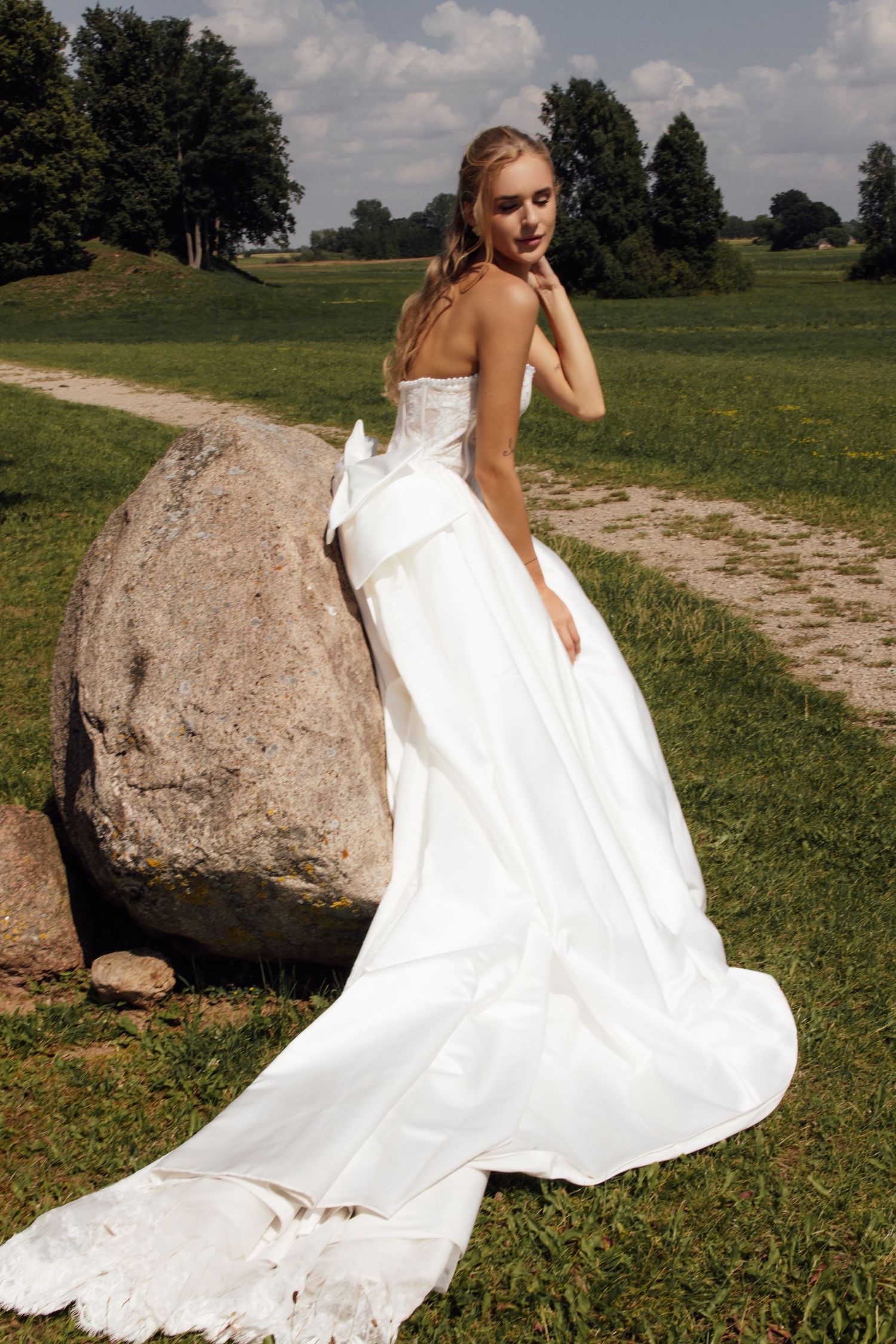 Bride in a strapless white ballgown with bow and train poses by a large rock in a green outdoor setting.