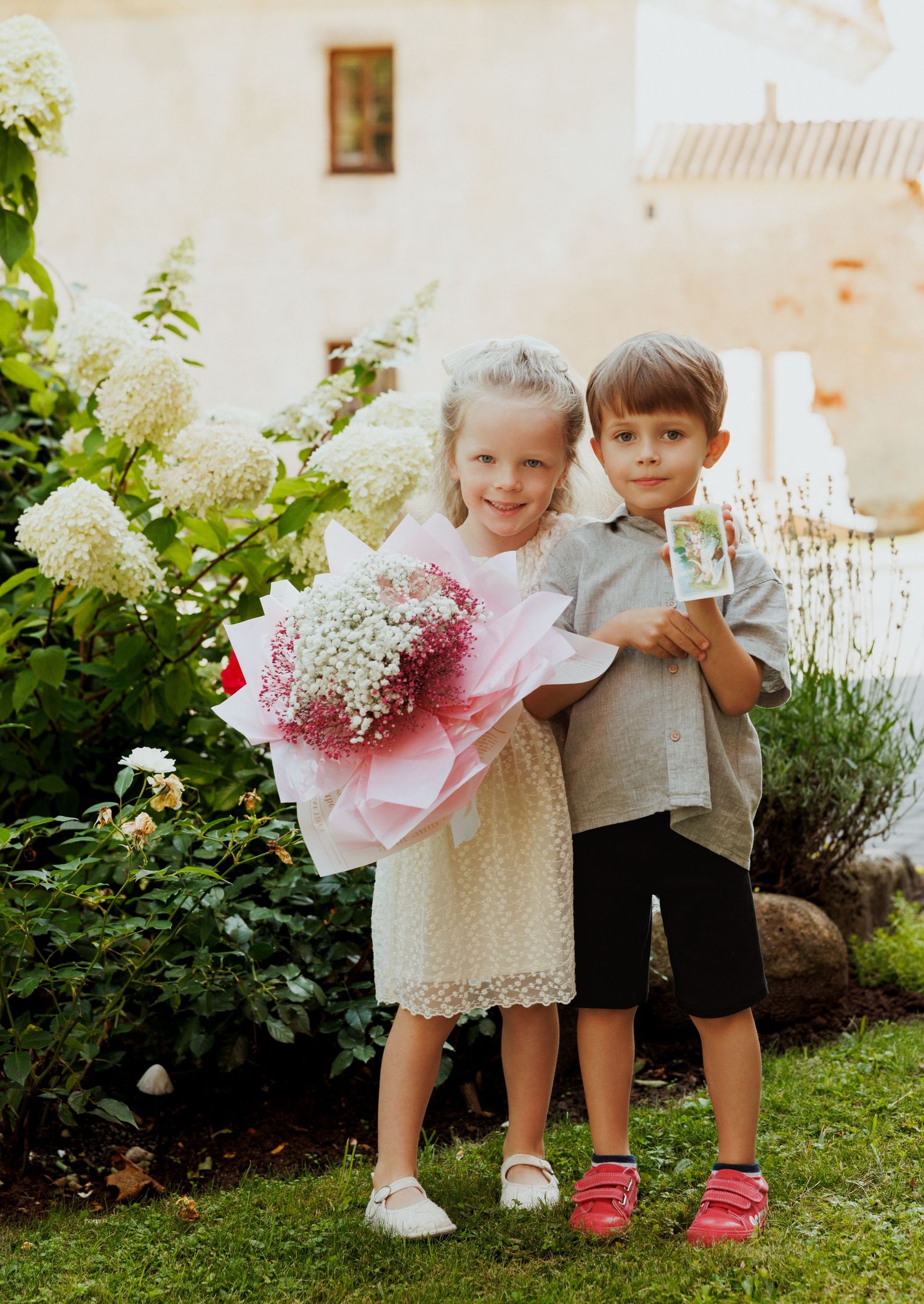 Two children standing in a garden holding floral bouquets against white hydrangea bushes.