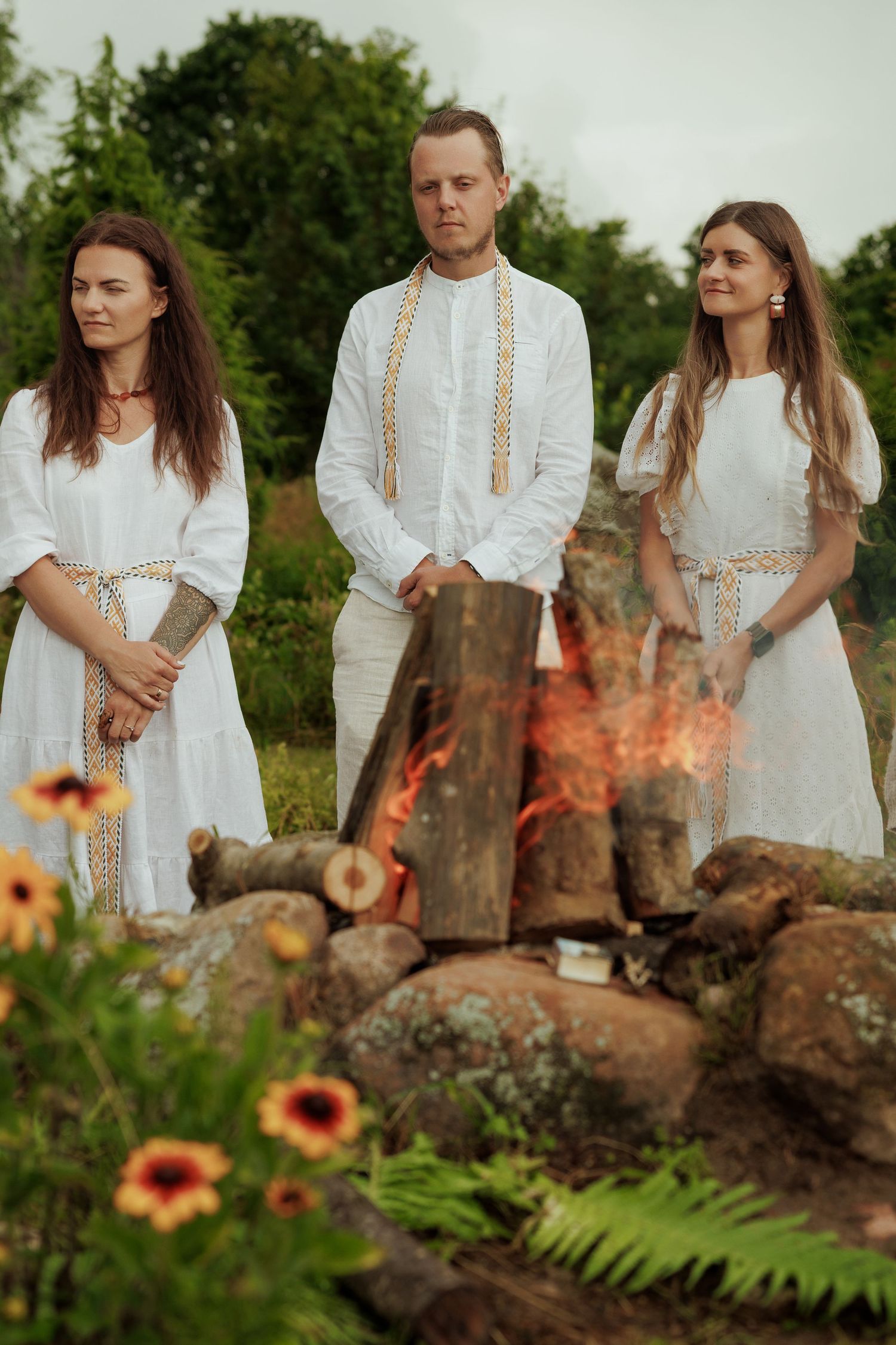 Three people in white traditional folk attire stand solemnly around a ceremonial bonfire surrounded by stones.