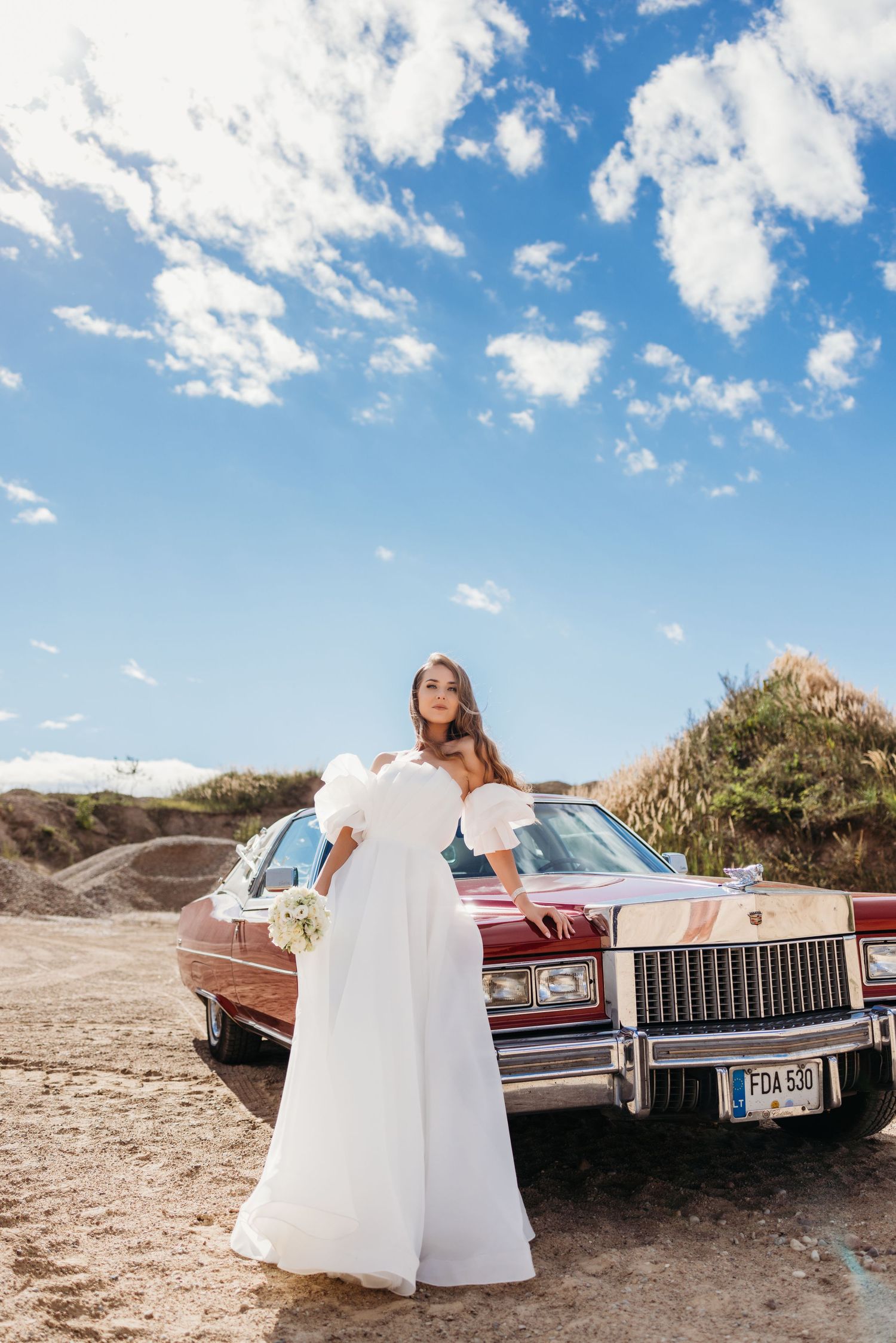 A vintage red Cadillac parked on a sandy beach under blue skies with white fluffy clouds.