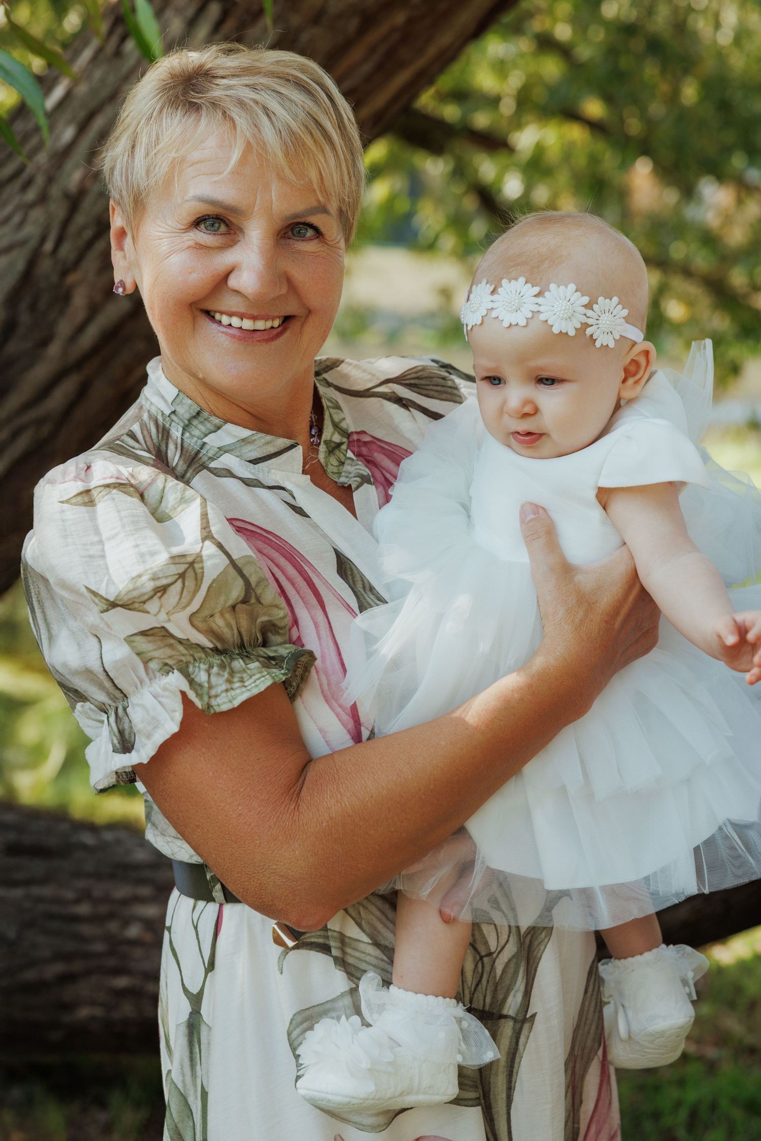 A tender moment captured outdoors showing an adult holding a baby wearing a white dress and floral headband.