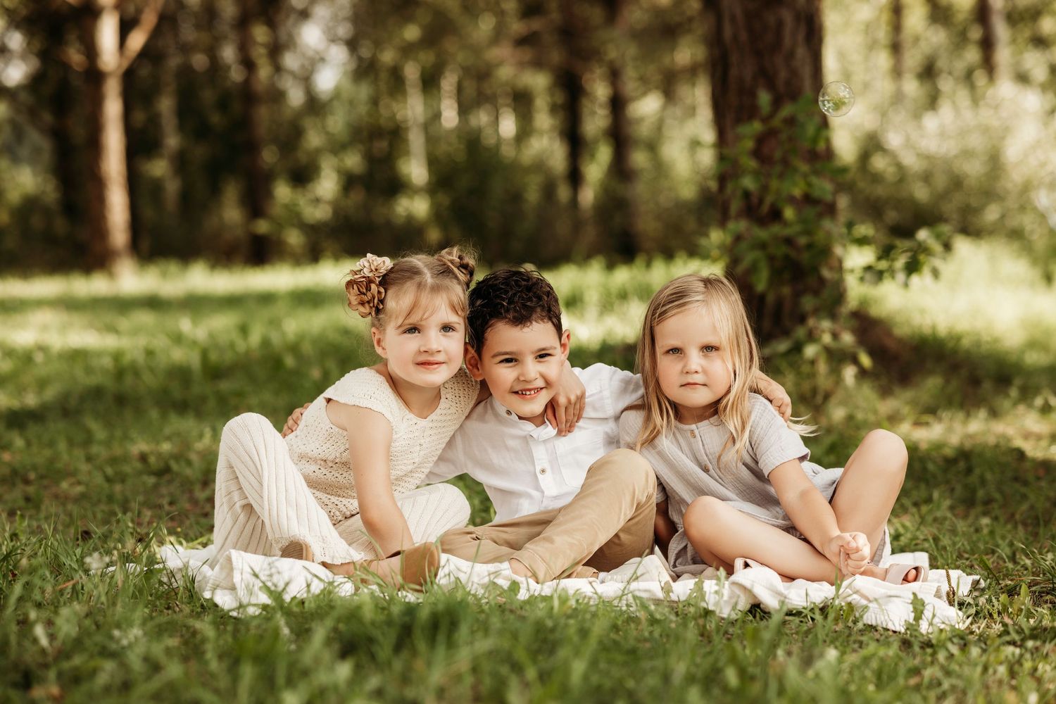 Three children in light colored clothing sitting together on blanket in sunny woodland setting.