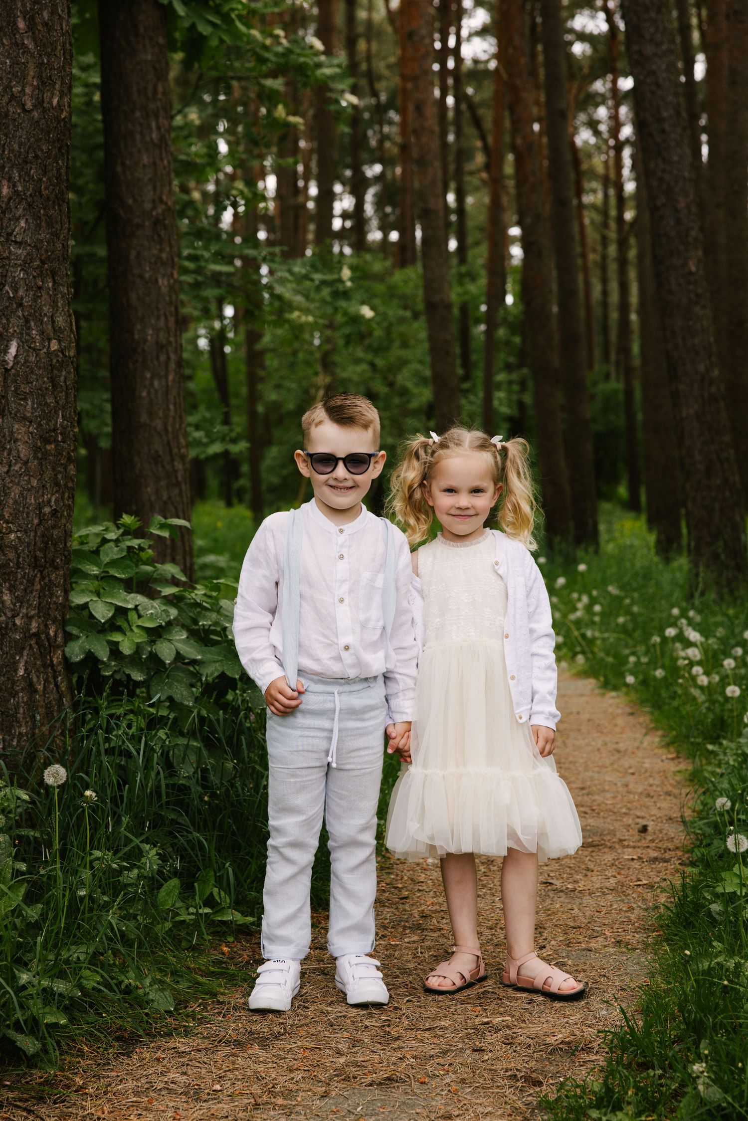 Two small children in formal white outfits standing on a forest path surrounded by greenery.