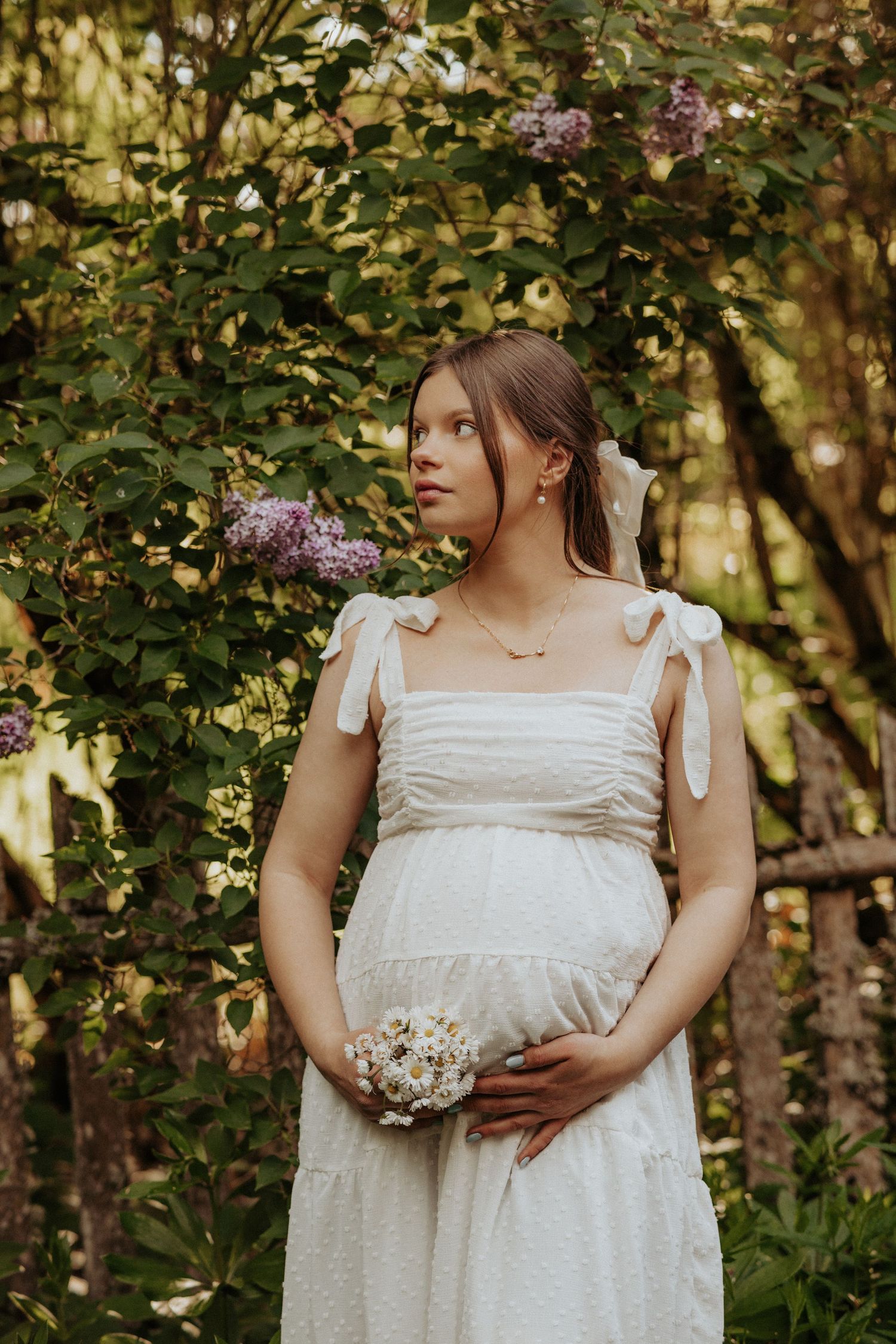 Ethereal maternity portrait in white dress among blooming lilac bushes.