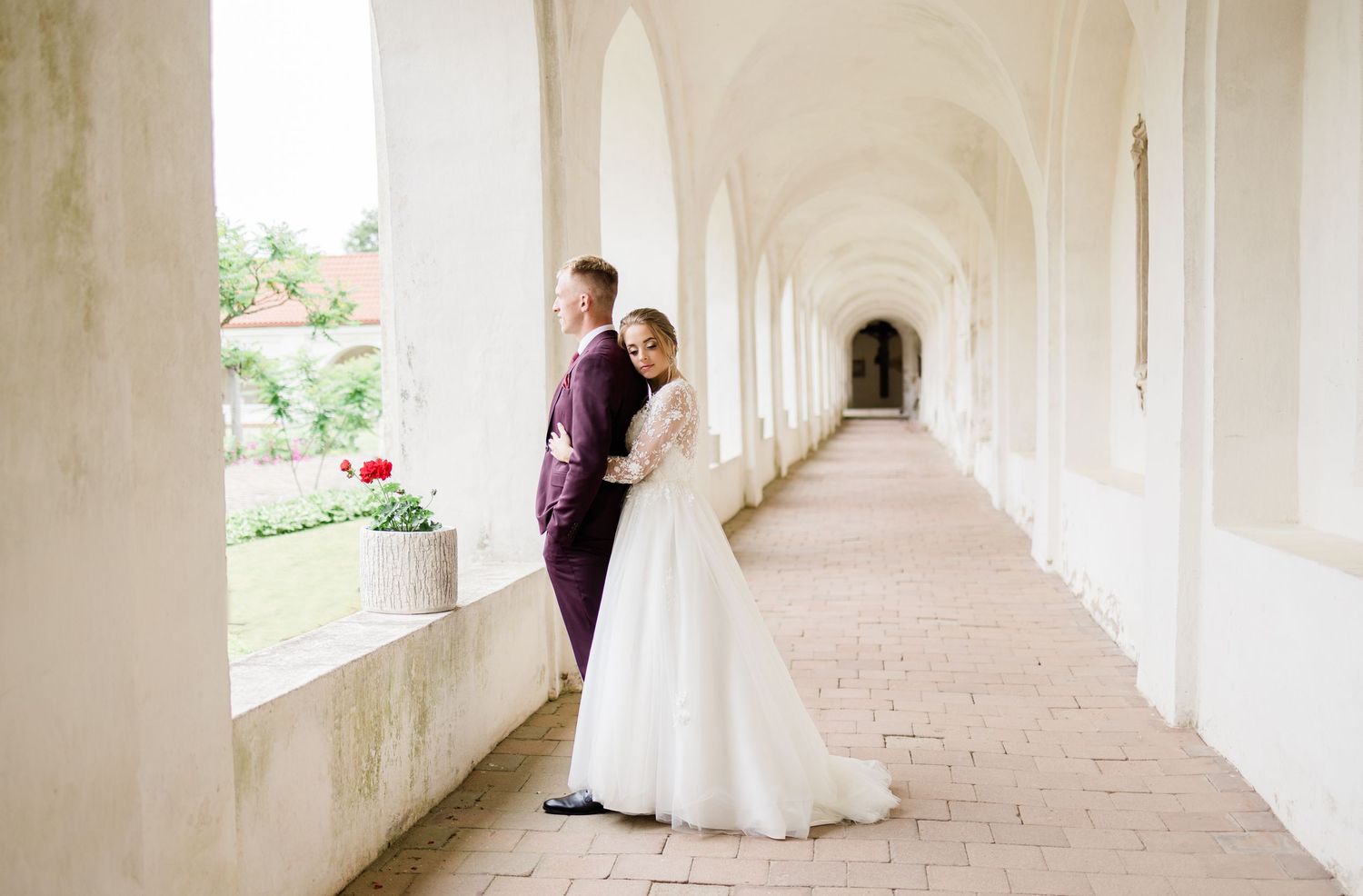Wedding couple shares intimate moment in elegant white colonnade hallway with arched windows.