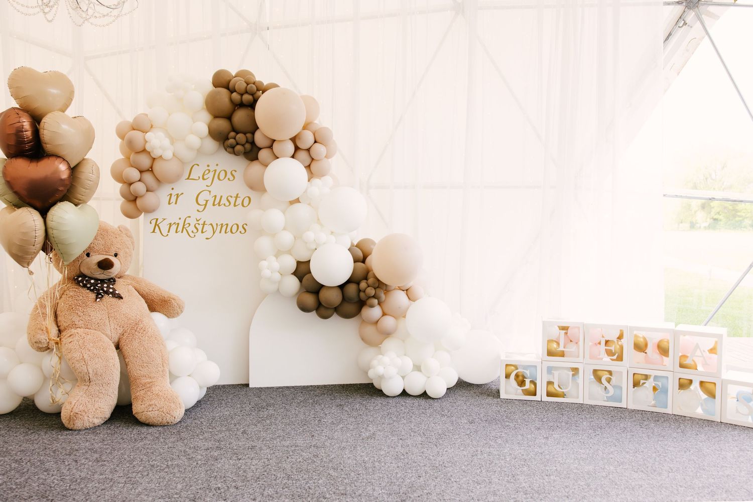 Baby shower backdrop with brown and white balloon arch and large teddy bear decorations.