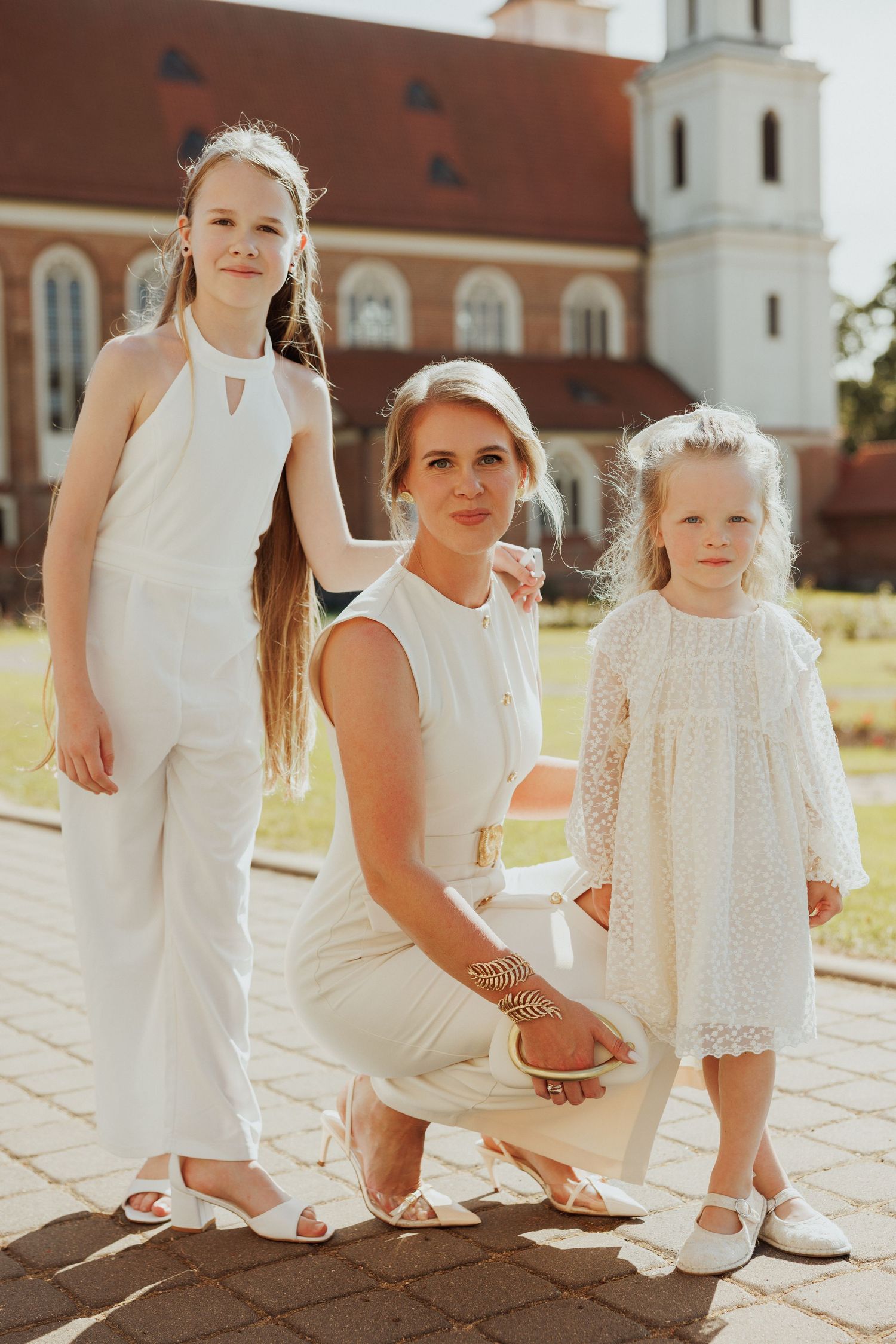 Three people in coordinated white outfits pose together in front of a church on a sunny day.