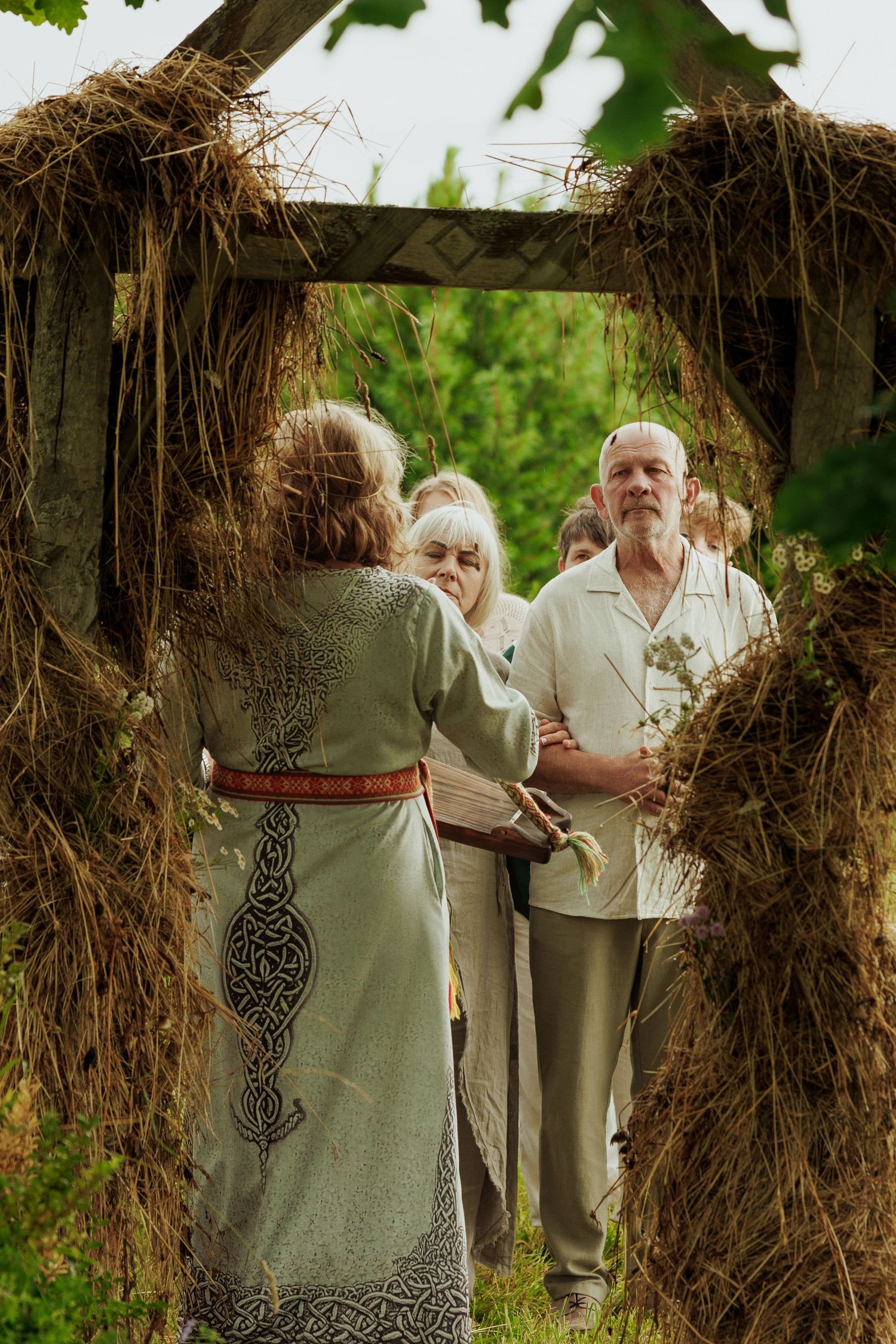 Elderly couple exchange vows outdoors under a rustic hay-covered arch during a traditional folk wedding ceremony.