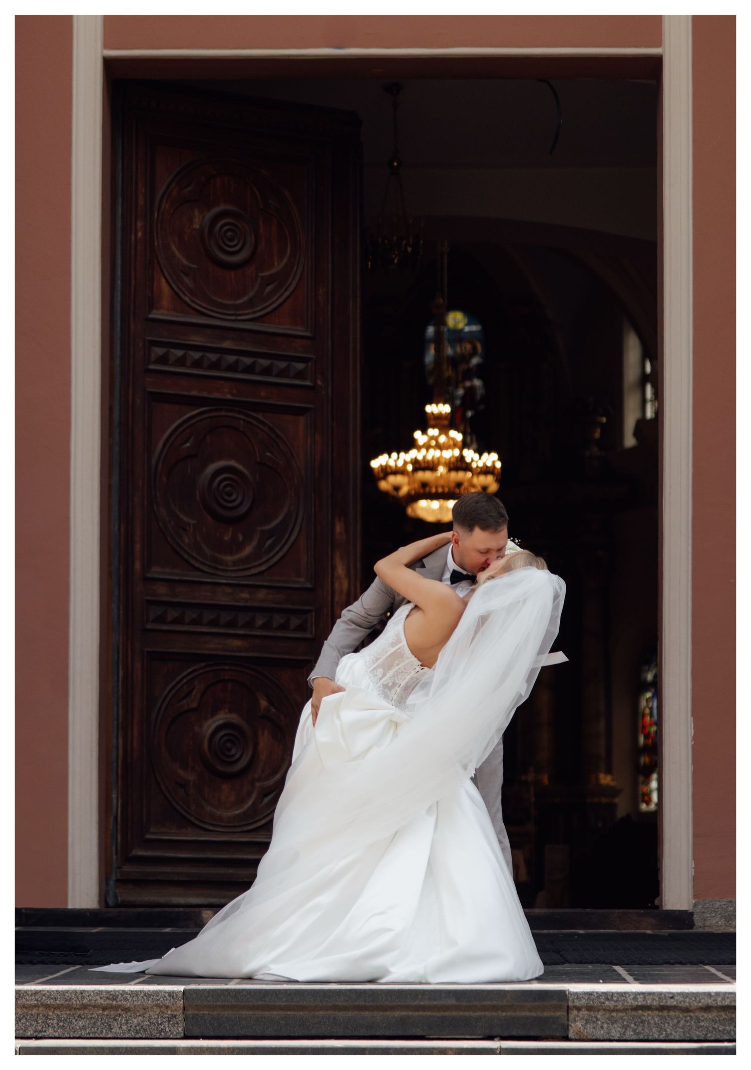 Bride and groom sharing a romantic dip kiss in front of ornate wooden church doors with chandelier backdrop.