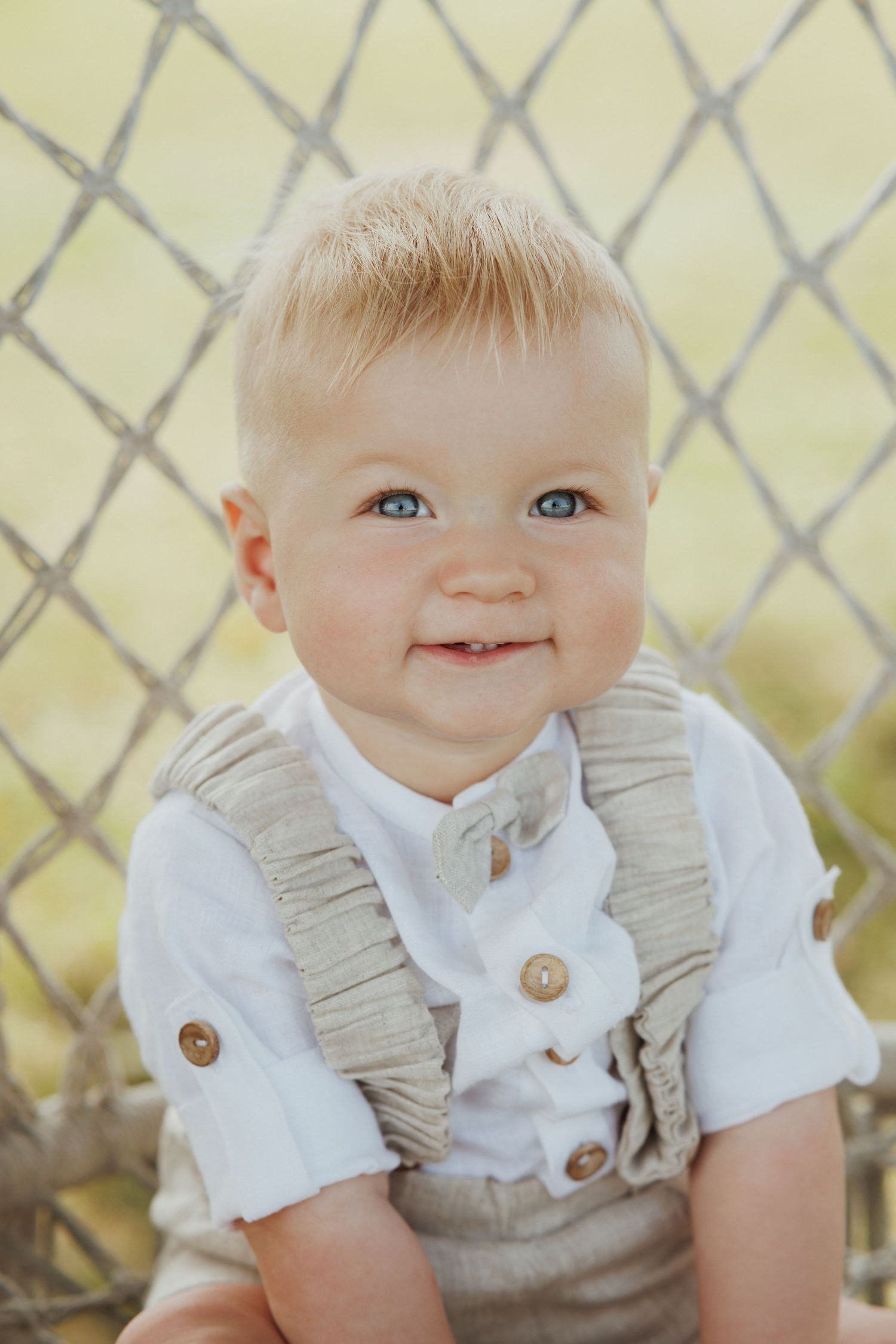 Small child wearing white shirt and beige vest sits against lattice background with a bright smile.