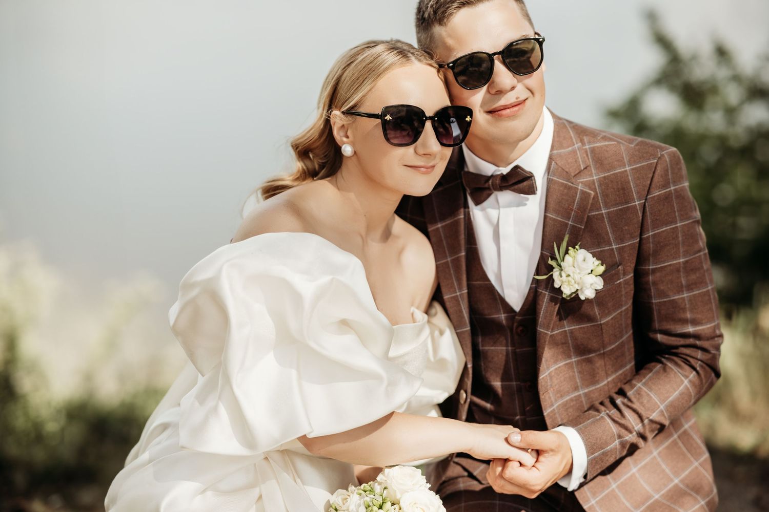 Stylish couple in wedding attire sharing a romantic moment outdoors wearing sunglasses.