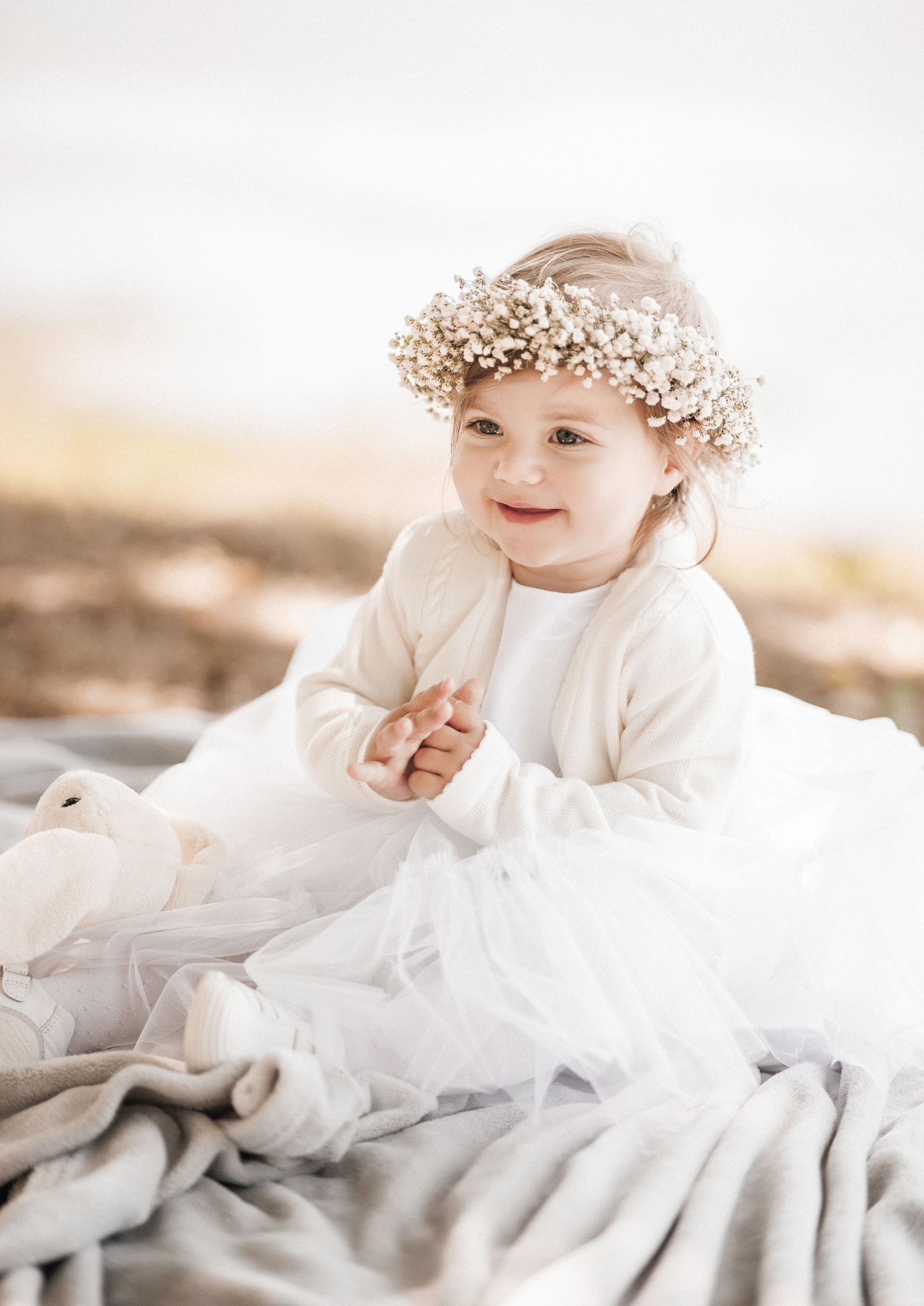 A baby wearing a white dress and floral crown lays on soft gray bedding in a dreamy portrait series.