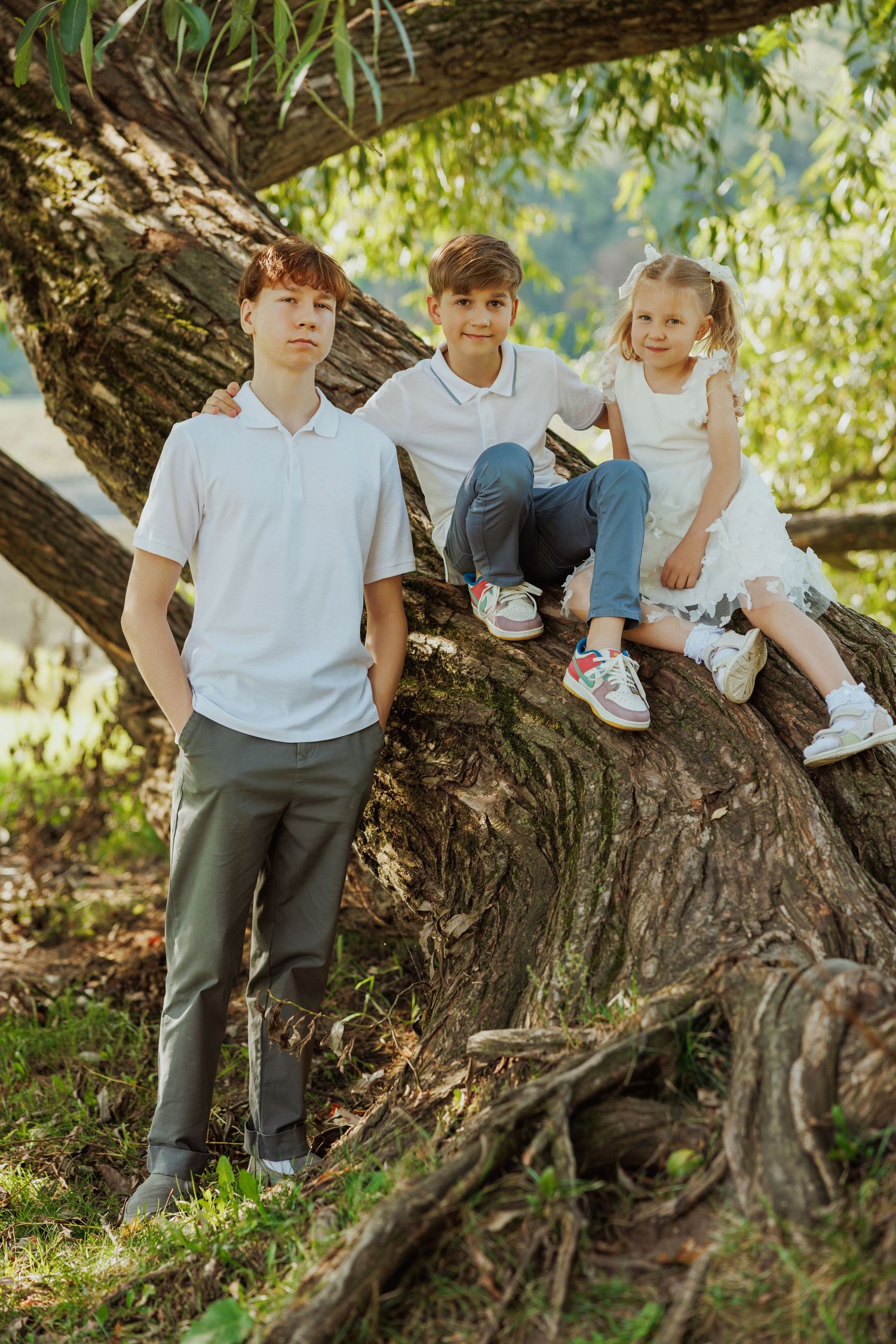Children in school uniforms sit together on large tree branches in a sunny garden setting.
