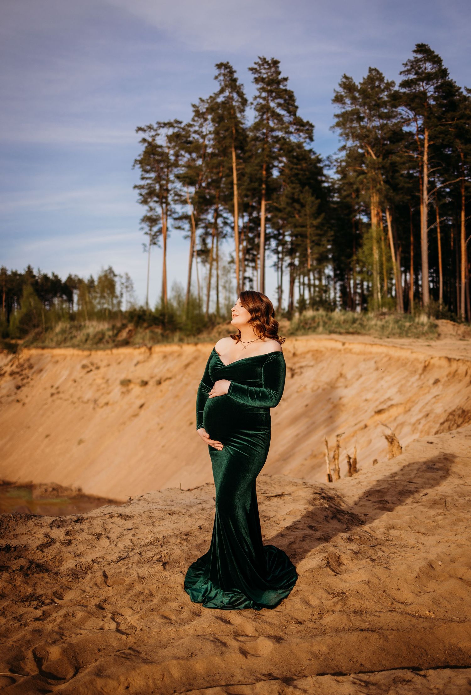 Dramatic maternity photos in emerald velvet dress against sandy dunes and pine trees.