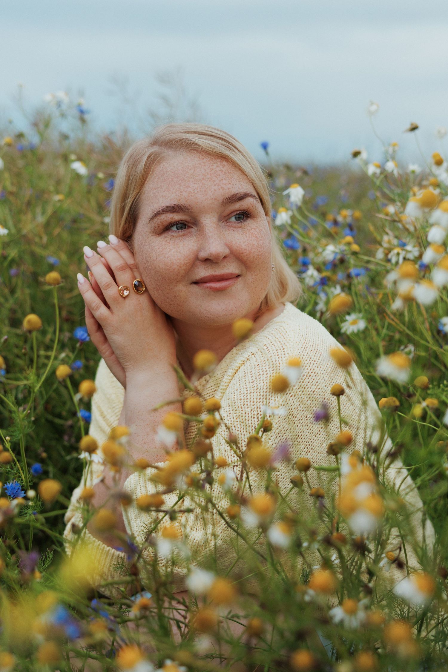 A smiling blonde woman sitting in a wildflower field with daisies and blue flowers on a cloudy day.