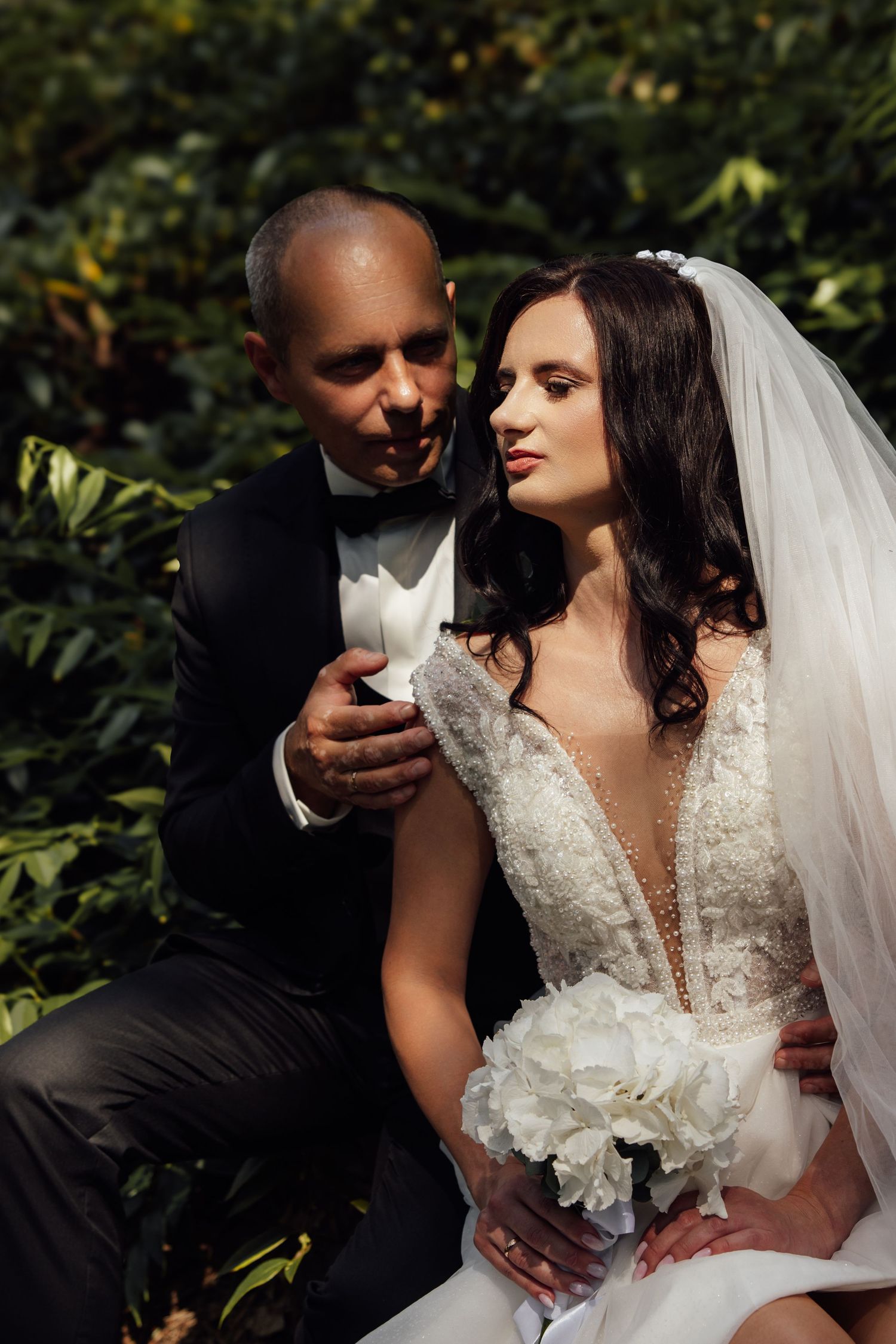 Groom in black tuxedo tenderly holds bride in lace wedding gown holding white peony bouquet amid green foliage.