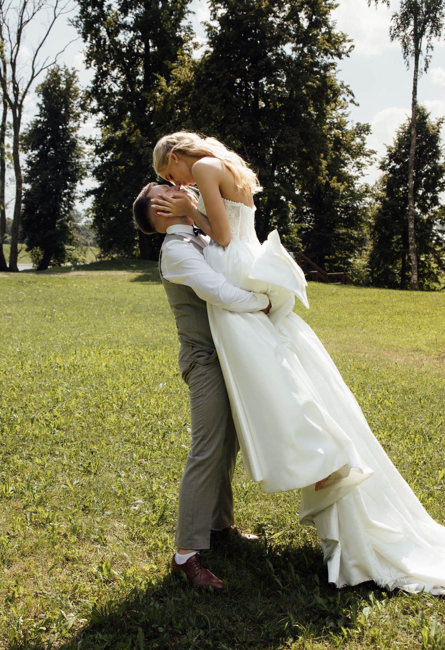 Bride and groom sharing a romantic kiss outdoors, groom lifting bride in white wedding dress on green lawn.