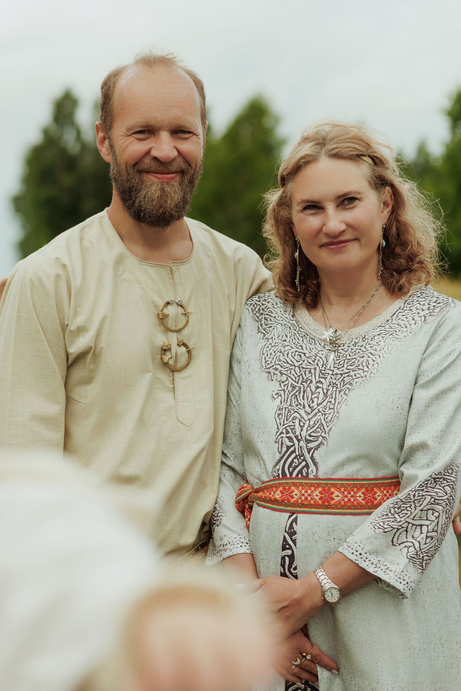Smiling couple in traditional folk costumes posing outdoors, man with beard in tan tunic, woman in gray embroidered dress.