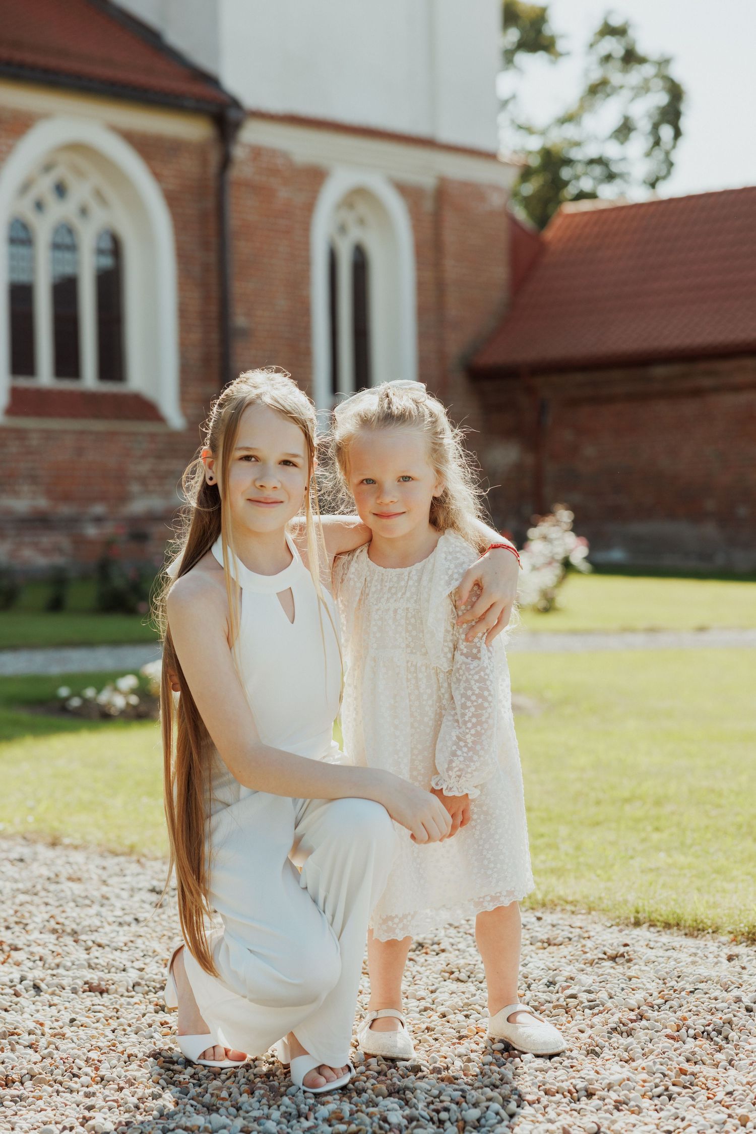 Two young girls in white dresses pose together on church grounds.