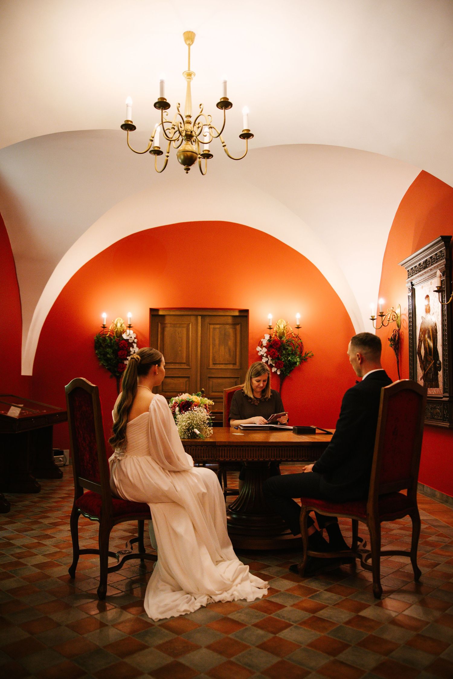 A wedding ceremony in an elegant red-walled room with arched ceilings and chandelier.