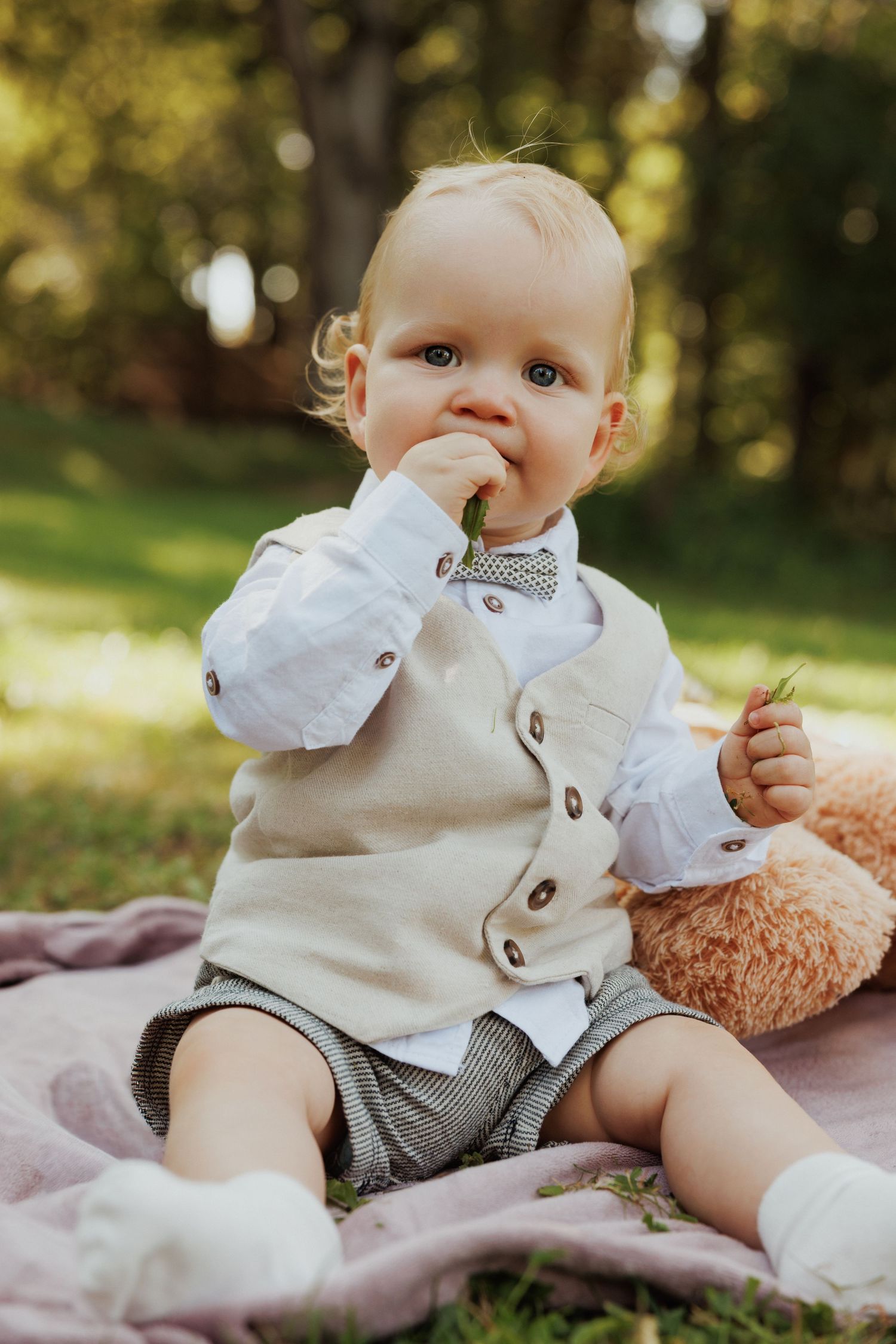 Baby in formal attire sits on a blanket outdoors while sucking thumb.