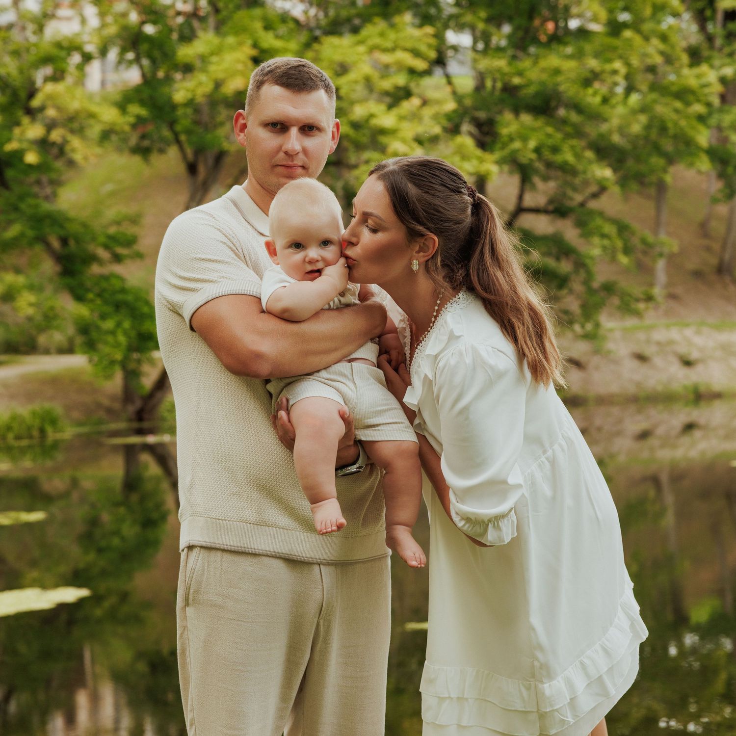 Family sharing tender moments by a tranquil lake with trees in the background during a sunny summer day.