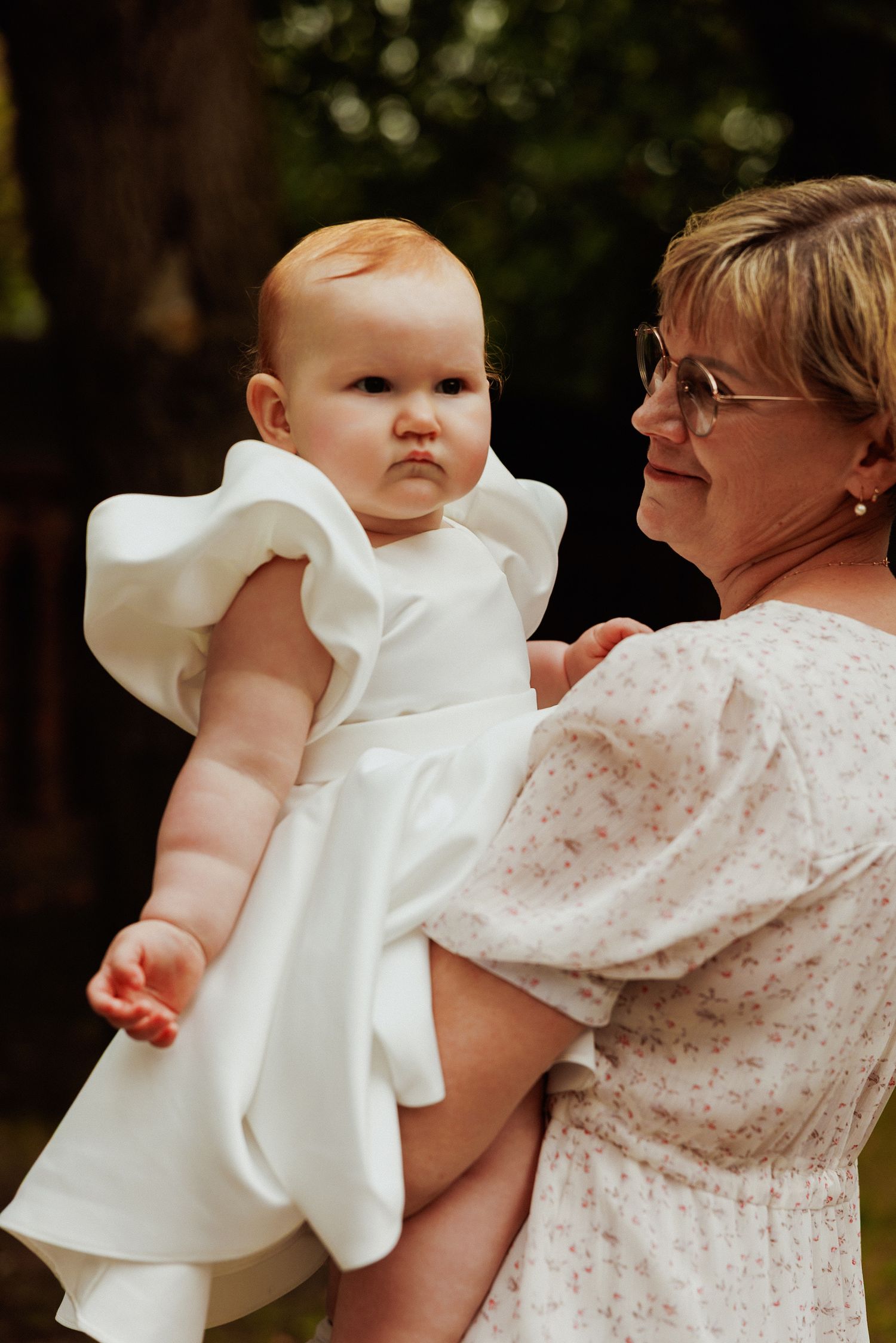 A baby wearing a white christening gown is held by someone in a floral print dress.