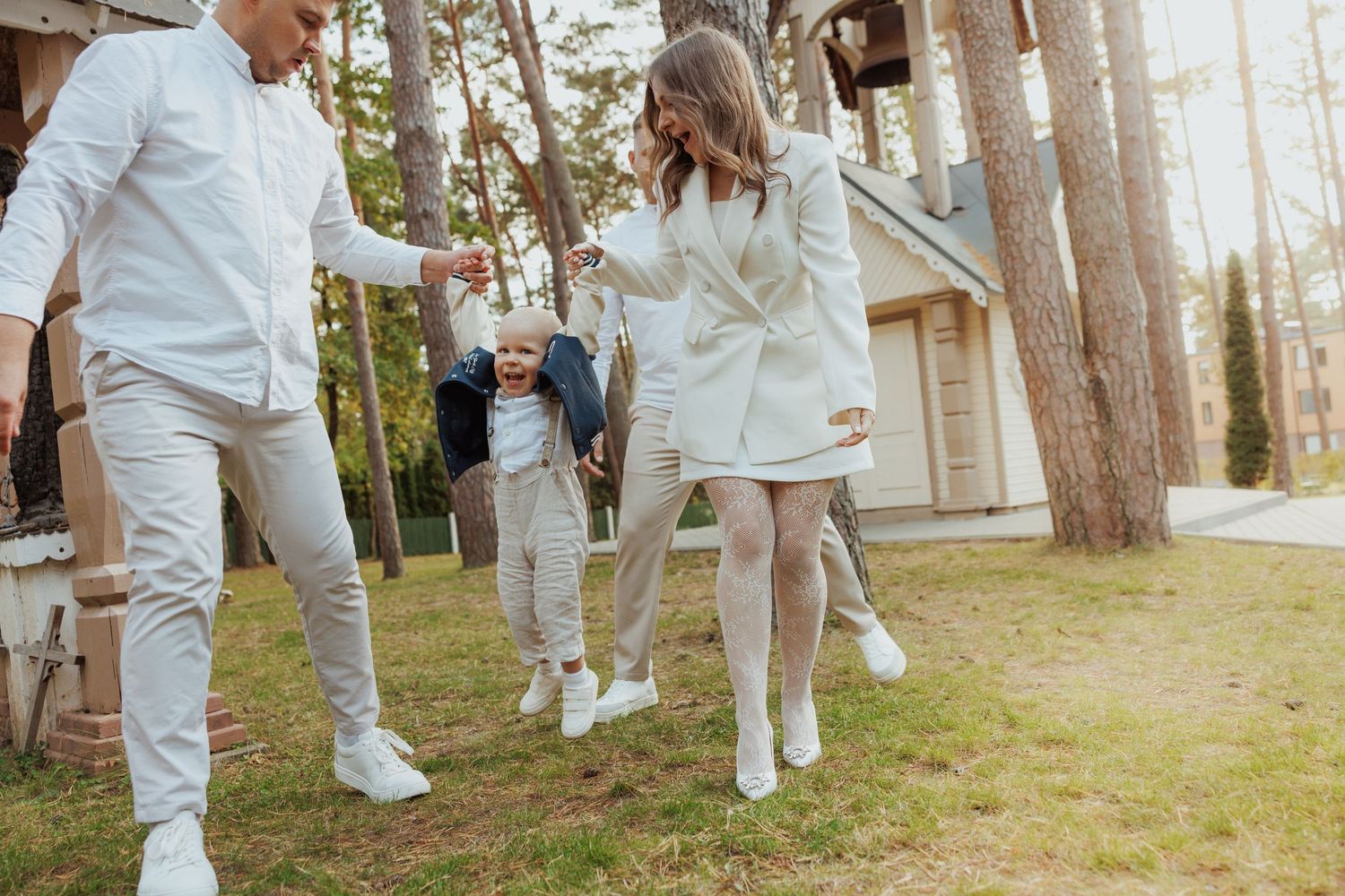 People in coordinated white outfits jump and play together on a grassy area near pine trees.