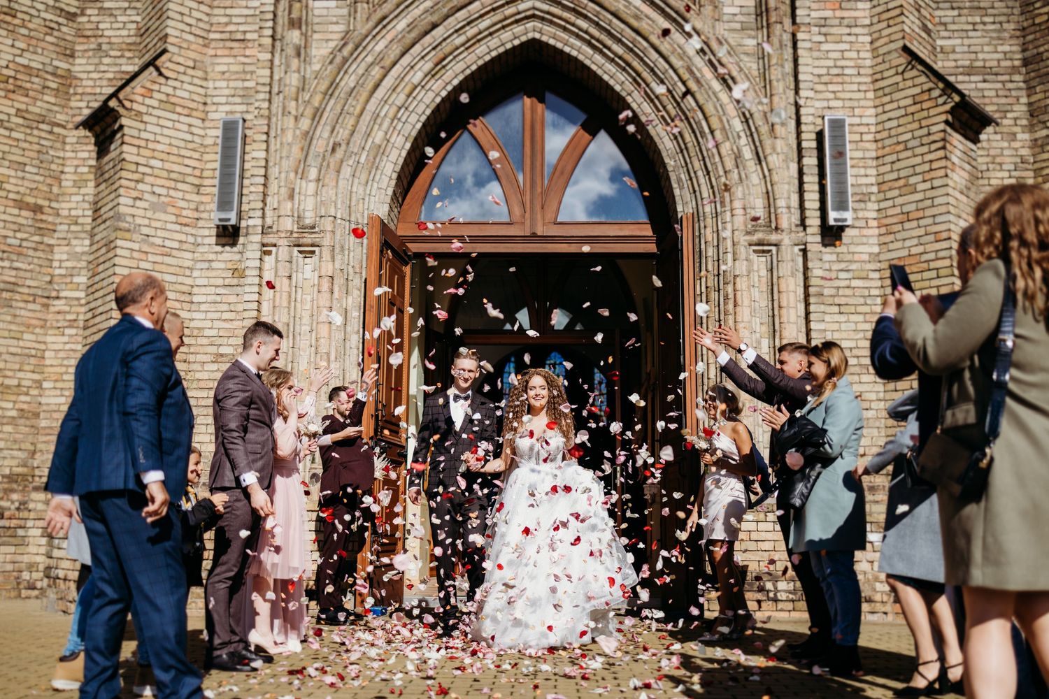 Wedding guests throw confetti outside historic stone church with arched doorway during joyful celebration.