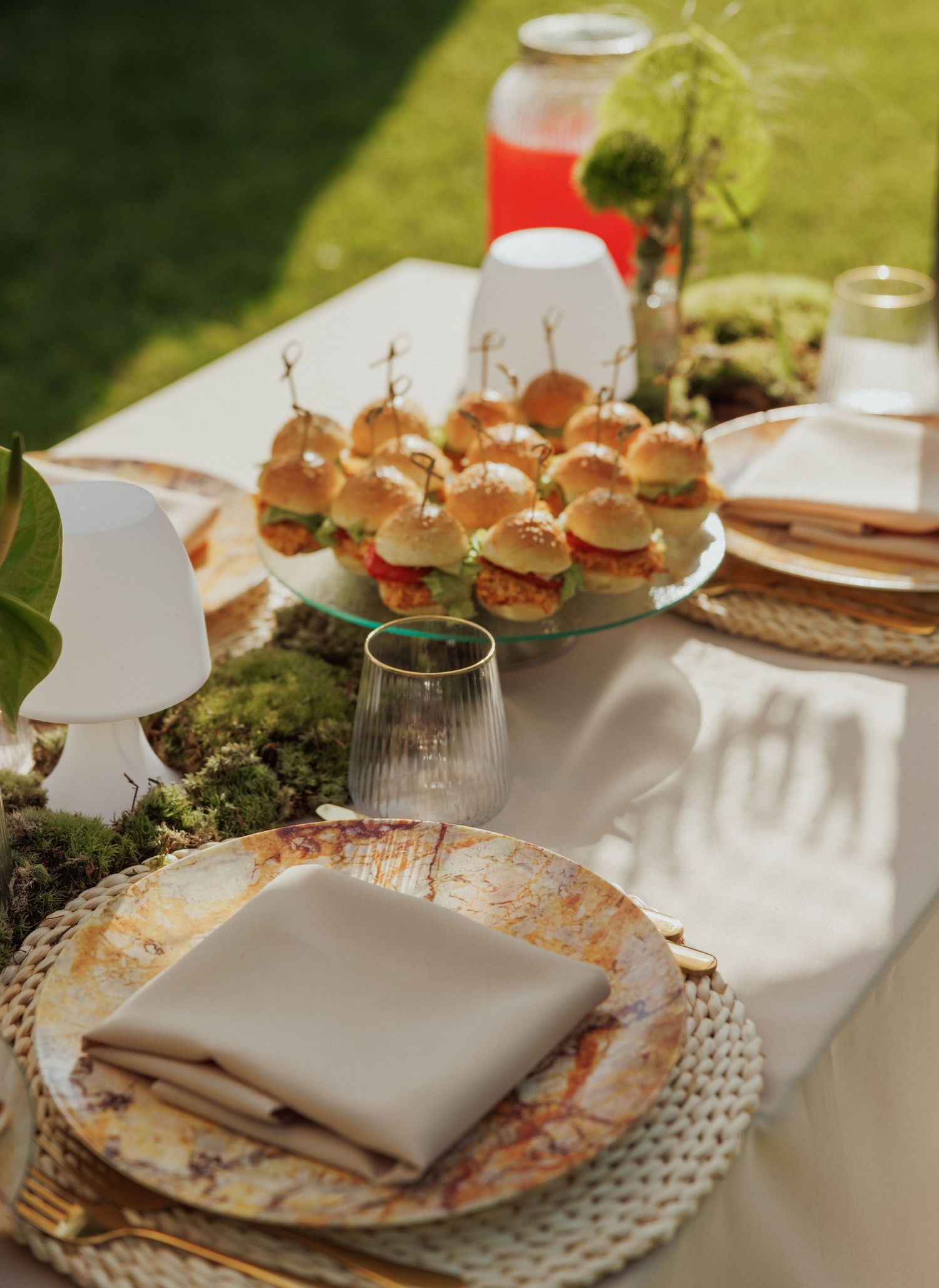 Elegant outdoor table setting with mini burgers and moss decor on textured plates.