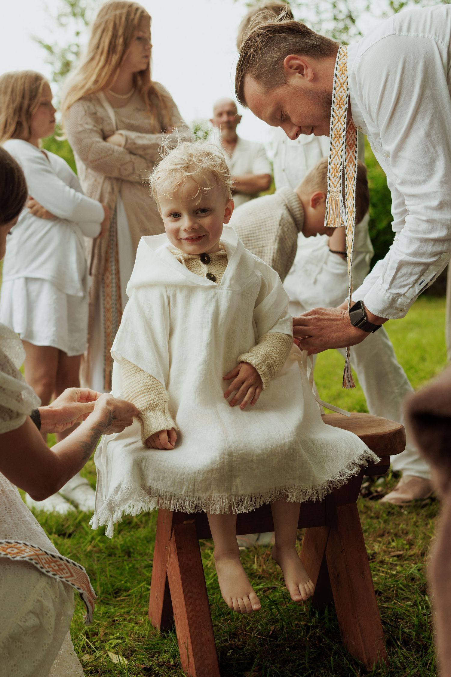 Smiling toddler in white linen outfit seated on wooden stool during outdoor folk ceremony.