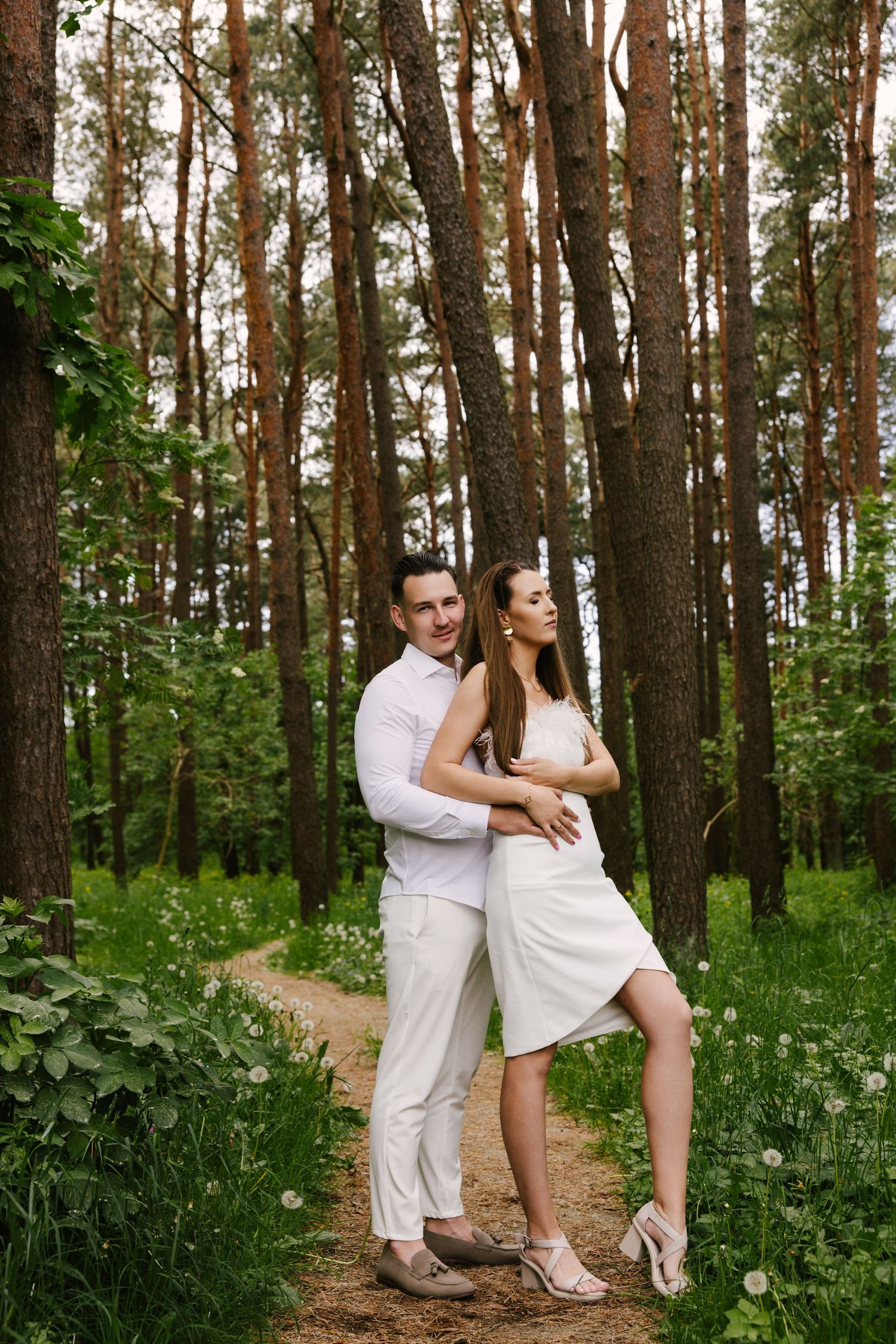 Romantic couple poses in matching white outfits along a forest path surrounded by tall pine trees.