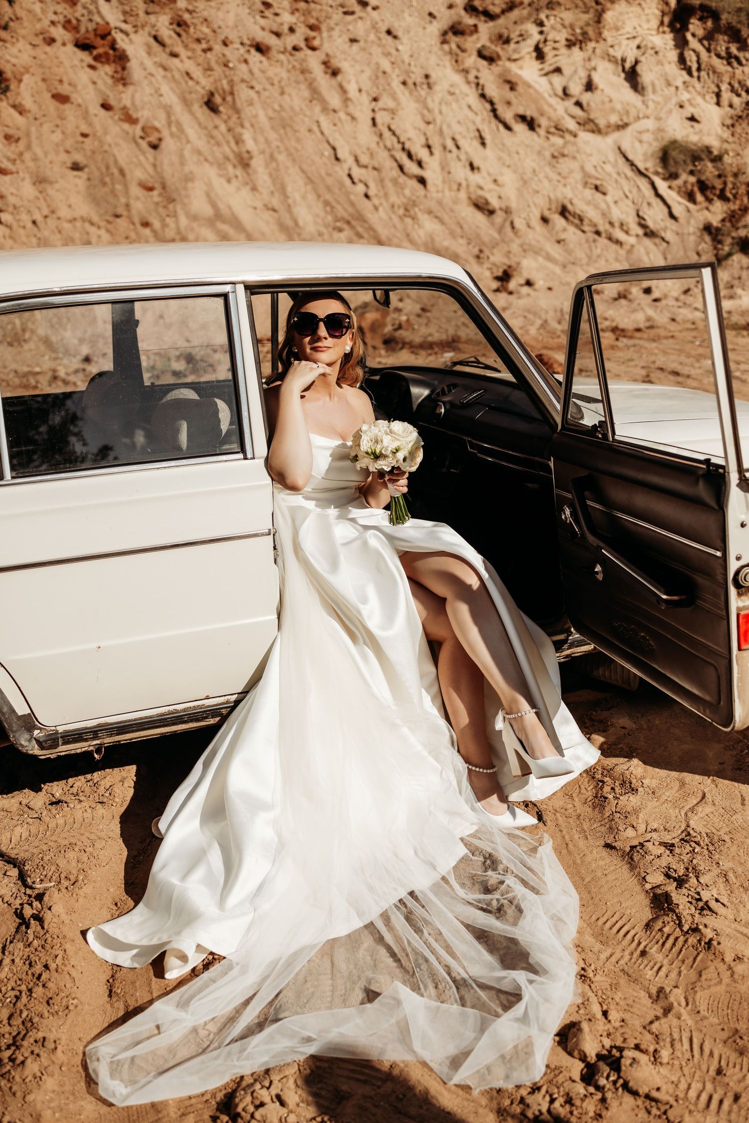 A flowing white wedding dress billows against a vintage white car parked on sandy terrain.