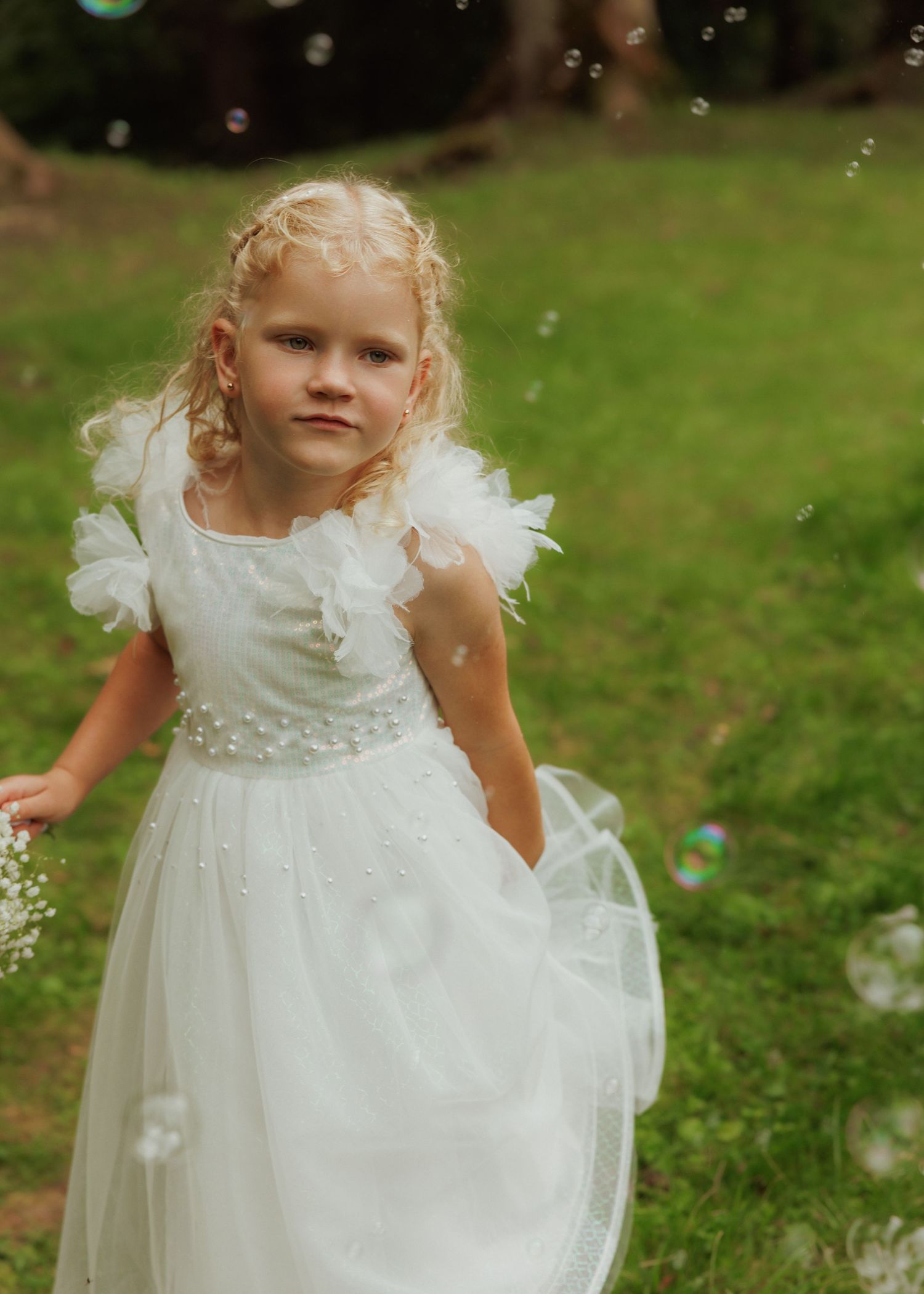 A young child in a white tulle dress stands on grass while soap bubbles float in the air around them.