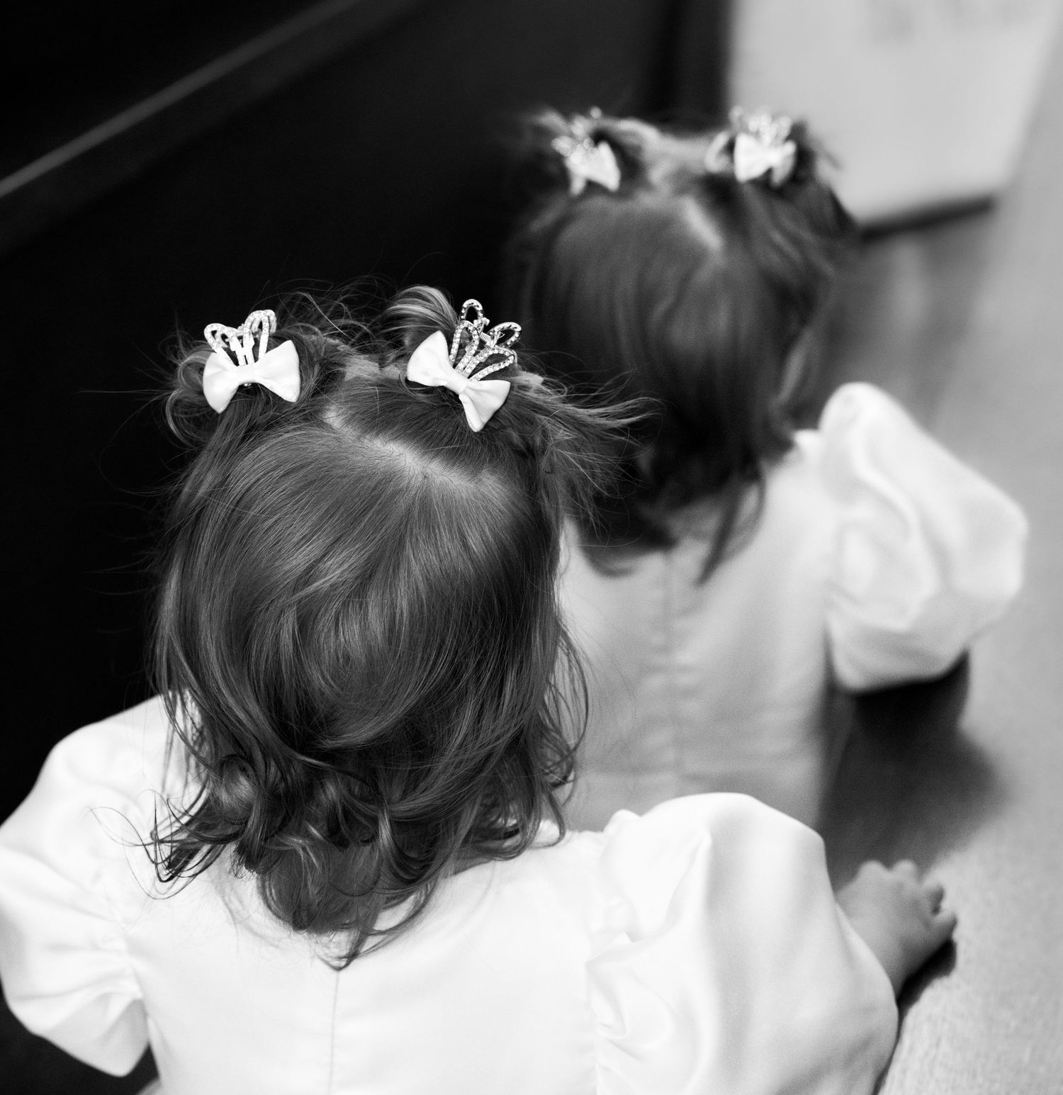 Two young children in white dresses with bows in their hair shown from behind in black and white photography.