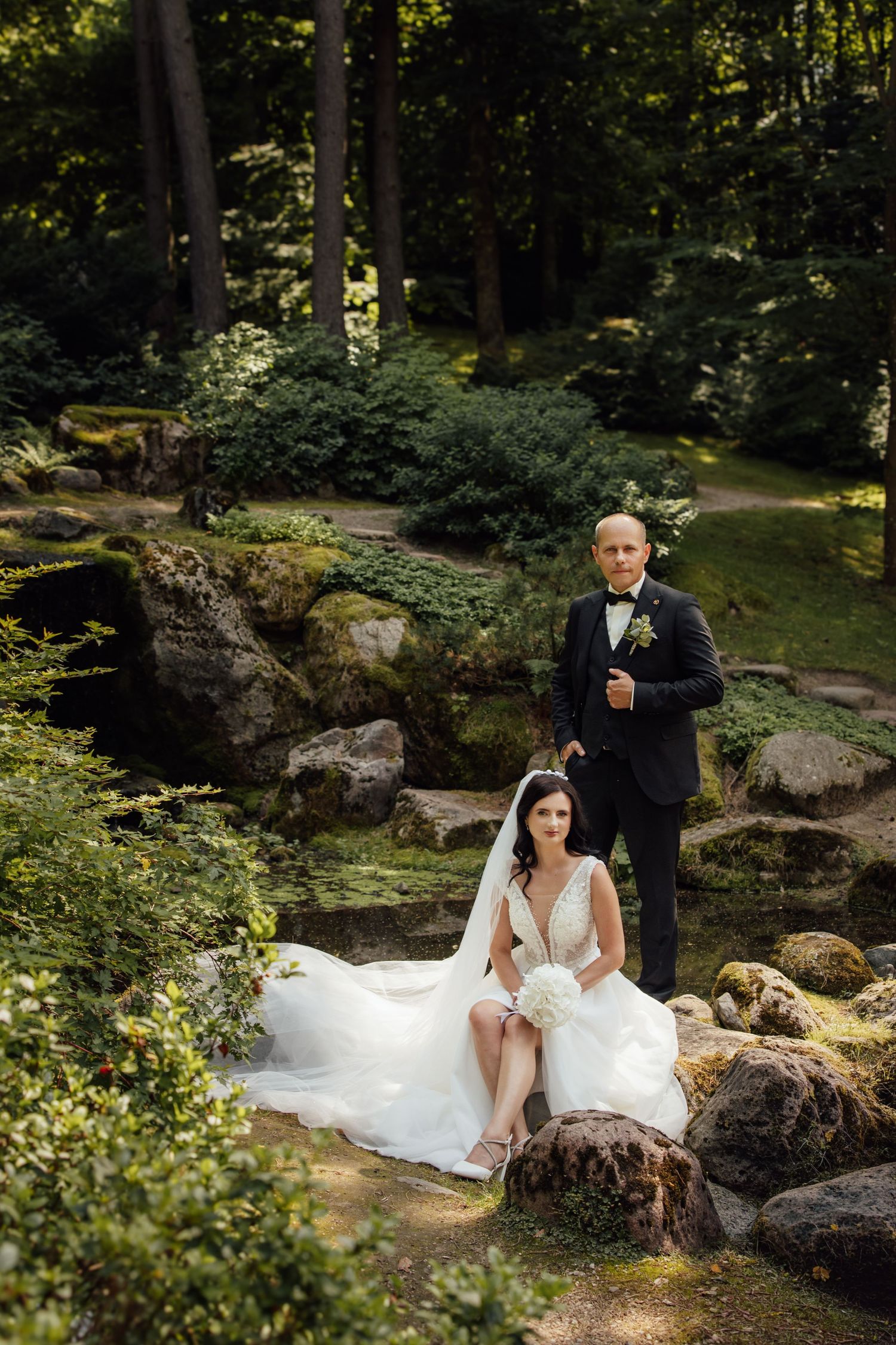 Bride and groom posing in a lush garden with rocks and greenery on their wedding day.