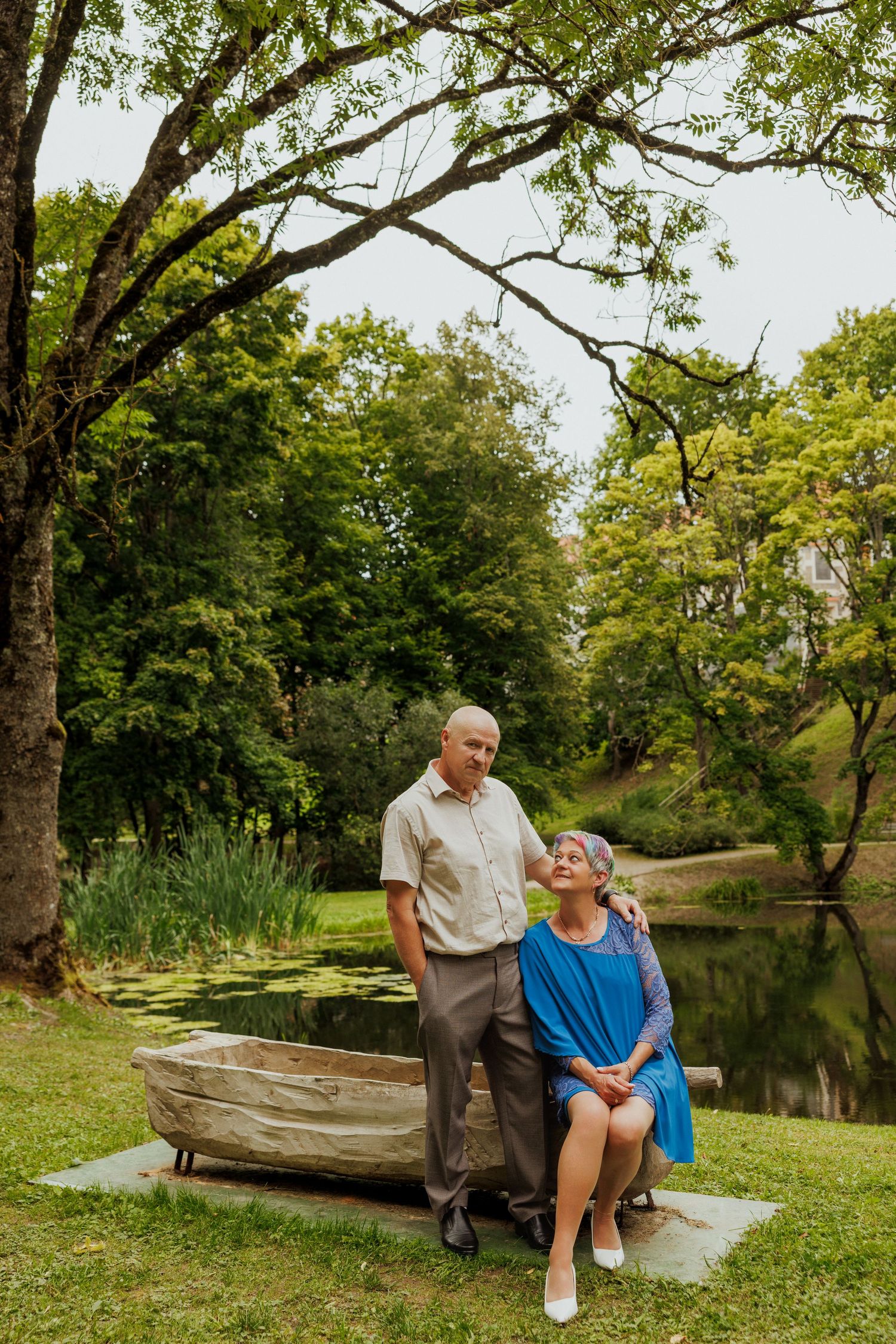 Couple sitting together on wooden boat by tranquil pond surrounded by lush green trees.