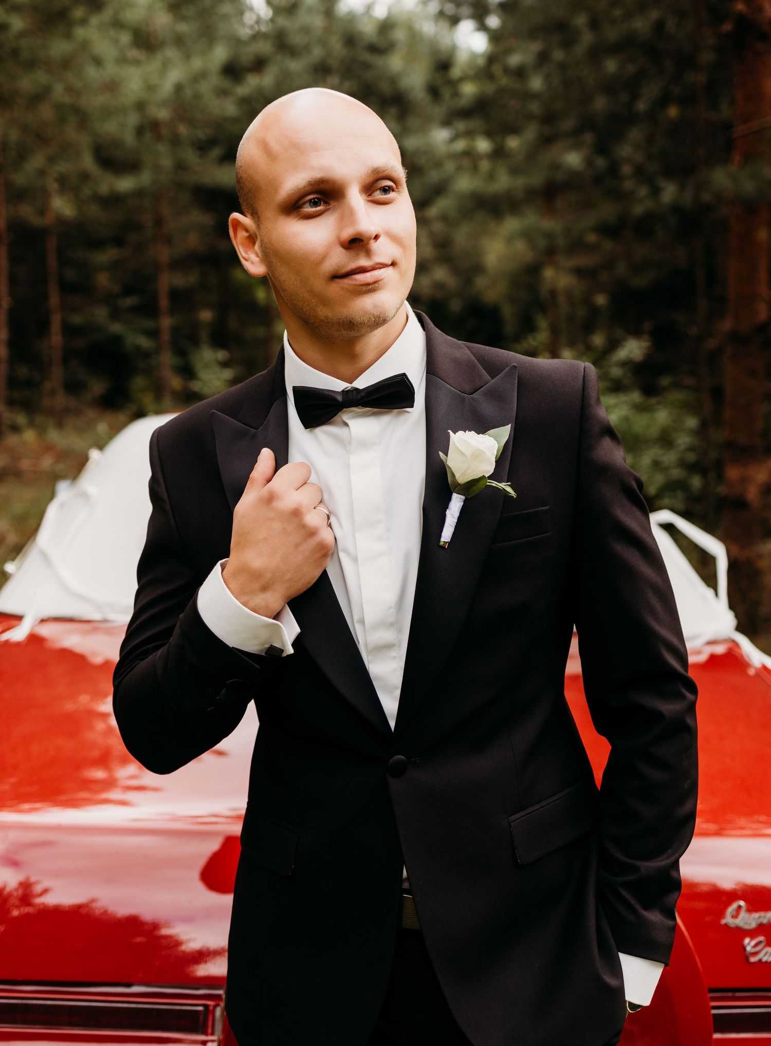 Groom in black tuxedo and bowtie poses confidently next to a classic red car.