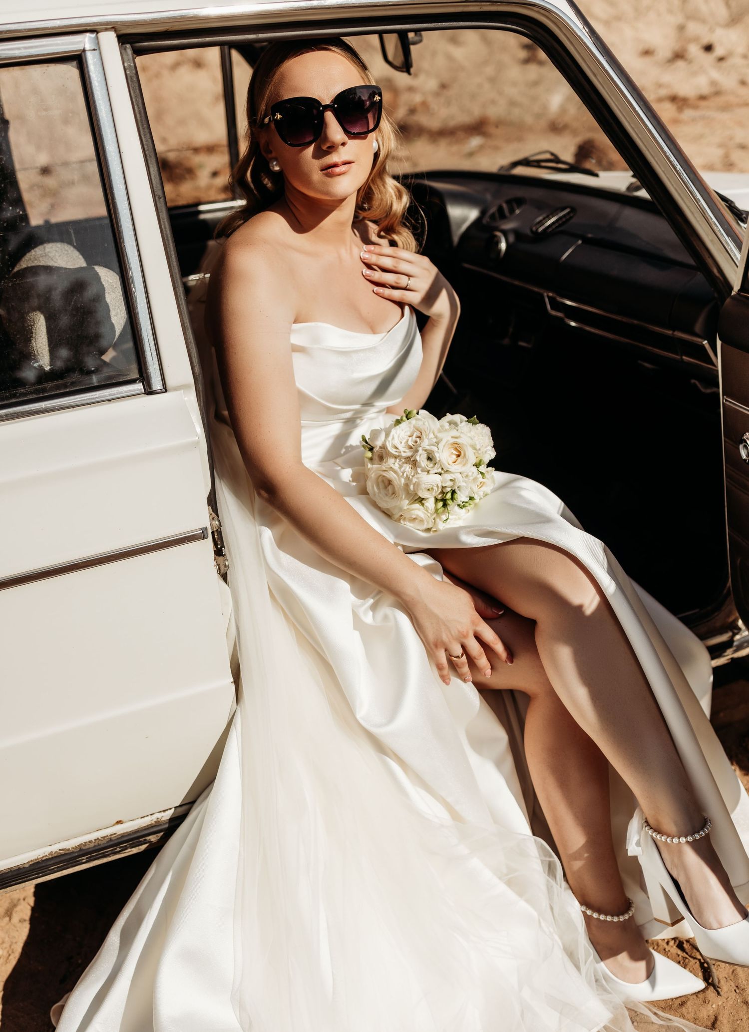 A bride in a white dress and sunglasses sits elegantly in a classic white vintage car.