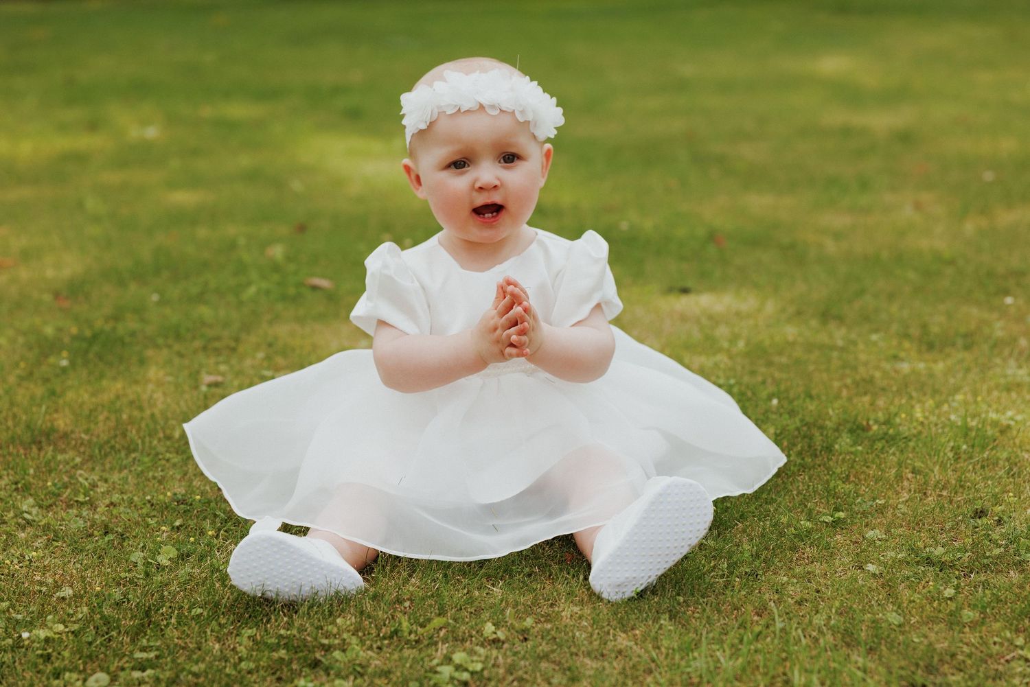 Baby in white christening gown sits and plays on green grass during outdoor photo session.