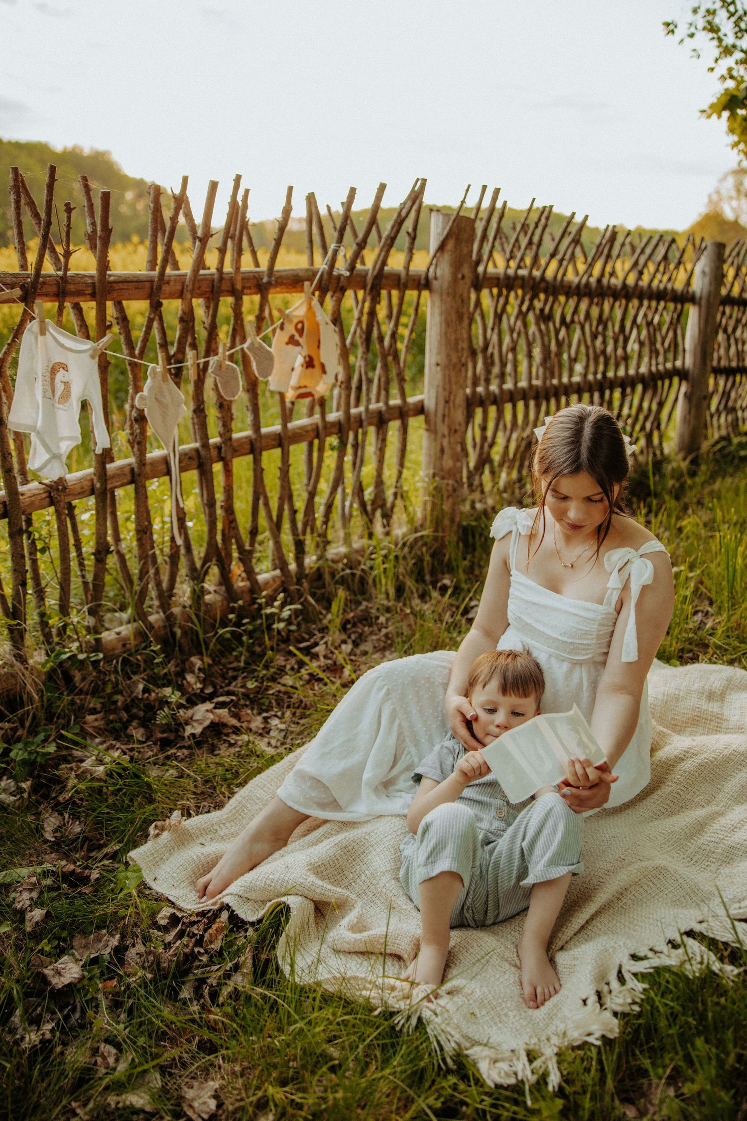 Sweet maternal moment on blanket by rustic fence with baby clothes hanging above.