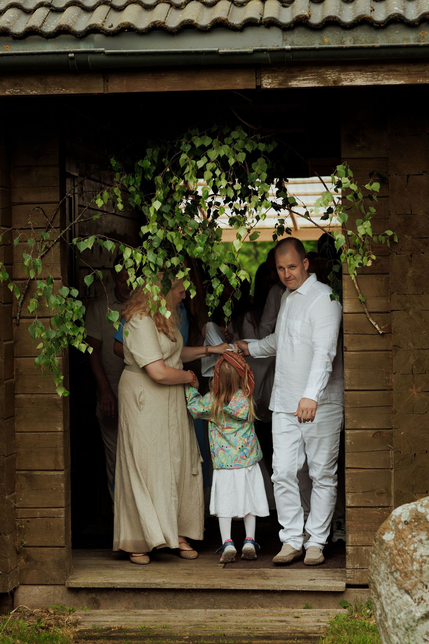 Family with young child standing in doorway of rustic wooden cabin decorated with birch branches.